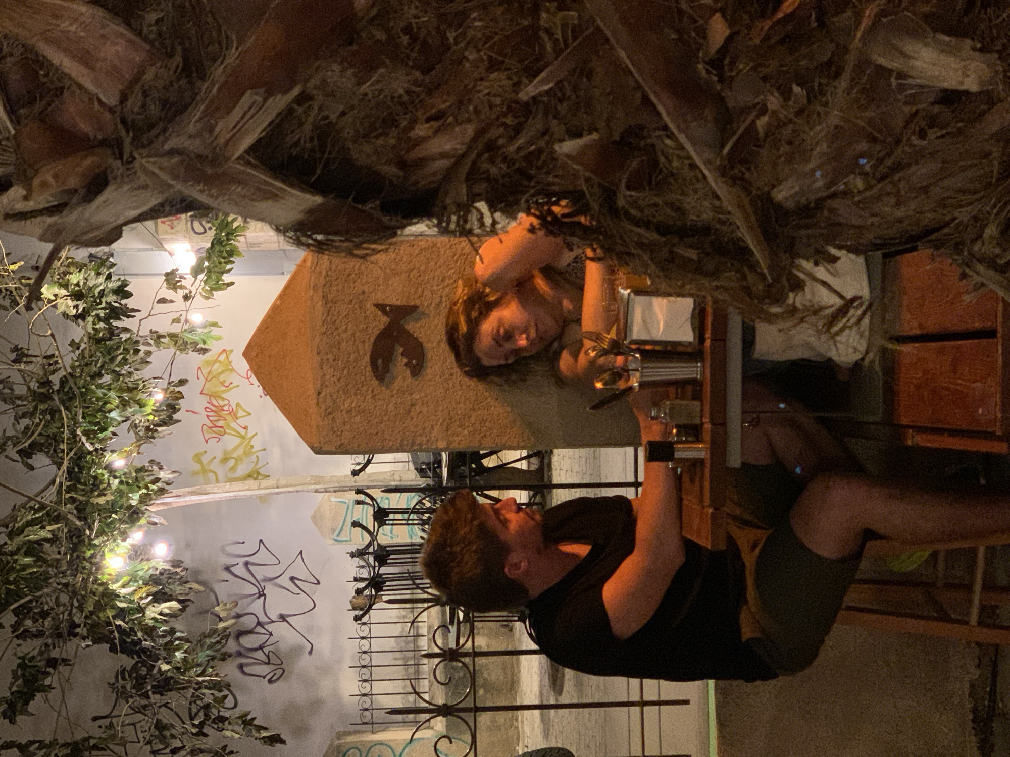 The image shows an outdoor setting in Tel Aviv, Israel, likely at night. Two individuals are present: a man is seated at a high table, wearing a black shirt and dark shorts, holding a glass or small container. A woman is standing nearby, also holding a glass. They appear to be at a bar or restaurant. A concrete pillar table with a crab symbol is visible, and behind the individuals is a wall with graffiti. A tree with lights is also present. The area is lit by artificial light, and the foreground contains foliage, likely from a palm tree.