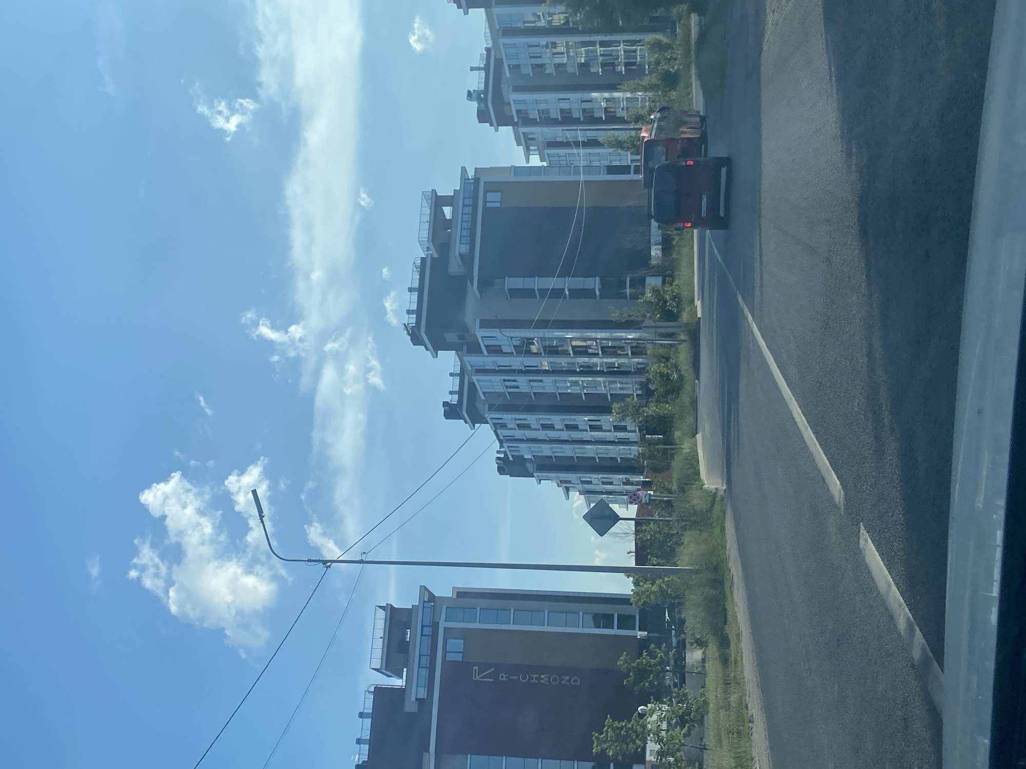 The image depicts a clear, sunny day with a blue sky and scattered white clouds, taken from a vehicle's perspective moving along a road. On the left side of the road, there are modern, multi-story apartment buildings with a dark brown facade and beige accents, featuring many windows. One building prominently displays the word "RICHMOND" vertically in white lettering, accompanied by a stylized "R" logo. To the right of these buildings, more apartment blocks are visible, characterized by a lighter color scheme with vertical lines and numerous balconies.
A street lamp with power lines stretches across the scene. The road itself is asphalt with visible white lane markings. In the middle ground, a red van and a red bus are traveling away from the camera. The surrounding area is somewhat undeveloped, with patches of grass, bushes, and young trees lining the road and around the buildings. A diamond-shaped traffic sign is visible on a pole. The overall impression is of a newly developed urban or suburban area, likely a residential complex. Based on visual cues from similar apartment building styles and the presence of the "RICHMOND" residential complex, this scene is likely located in the city of Kazan, Russia, possibly in an area with new construction.