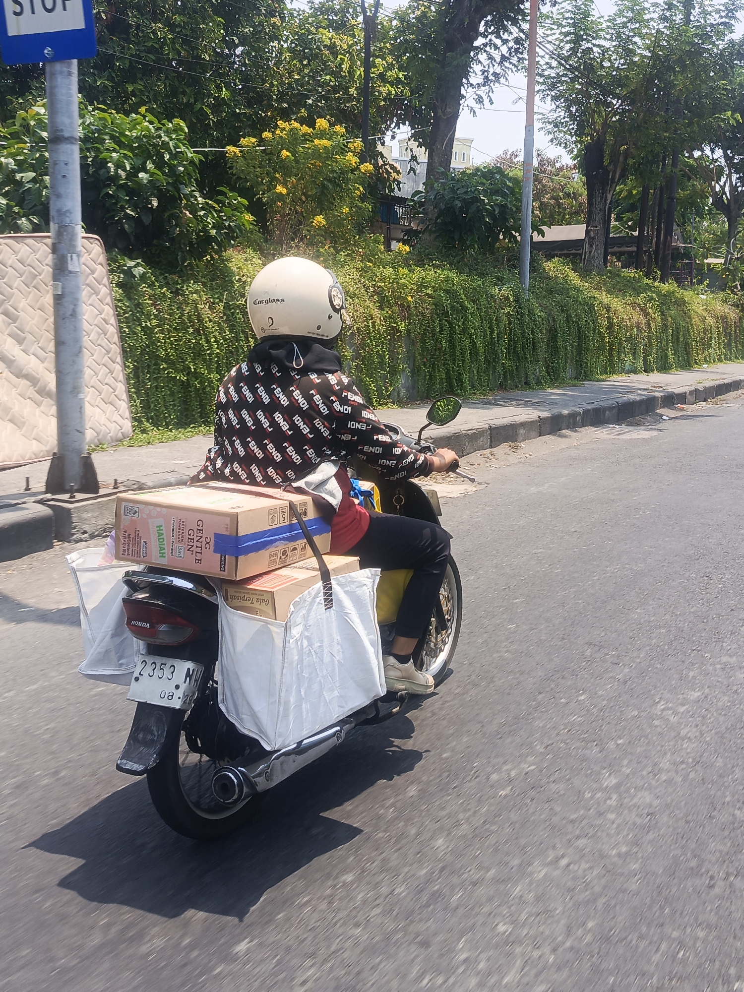 In a sunlit urban scene in Surabaya, Indonesia, a person rides a motorcycle loaded with parcels. The rider, wearing a white helmet with "Cargloss" branding and a black jacket adorned with a repeating "FENDI" pattern, is navigating a paved road. The motorcycle, a black Honda, has two large white bags strapped to the rear, and two cardboard boxes are stacked on top of them. One box is labeled "GENTLE GEN" and "HADIAH." A mattress is visible on the left side of the frame, leaning against a pole that bears a blue "STOP" sign. Lush green foliage and trees line the street, with some yellow flowers blooming amidst the greenery. The road surface is asphalt, and the motorcycle casts a distinct shadow. The license plate of the motorcycle reads "2353 NH" with "08.20" below it.