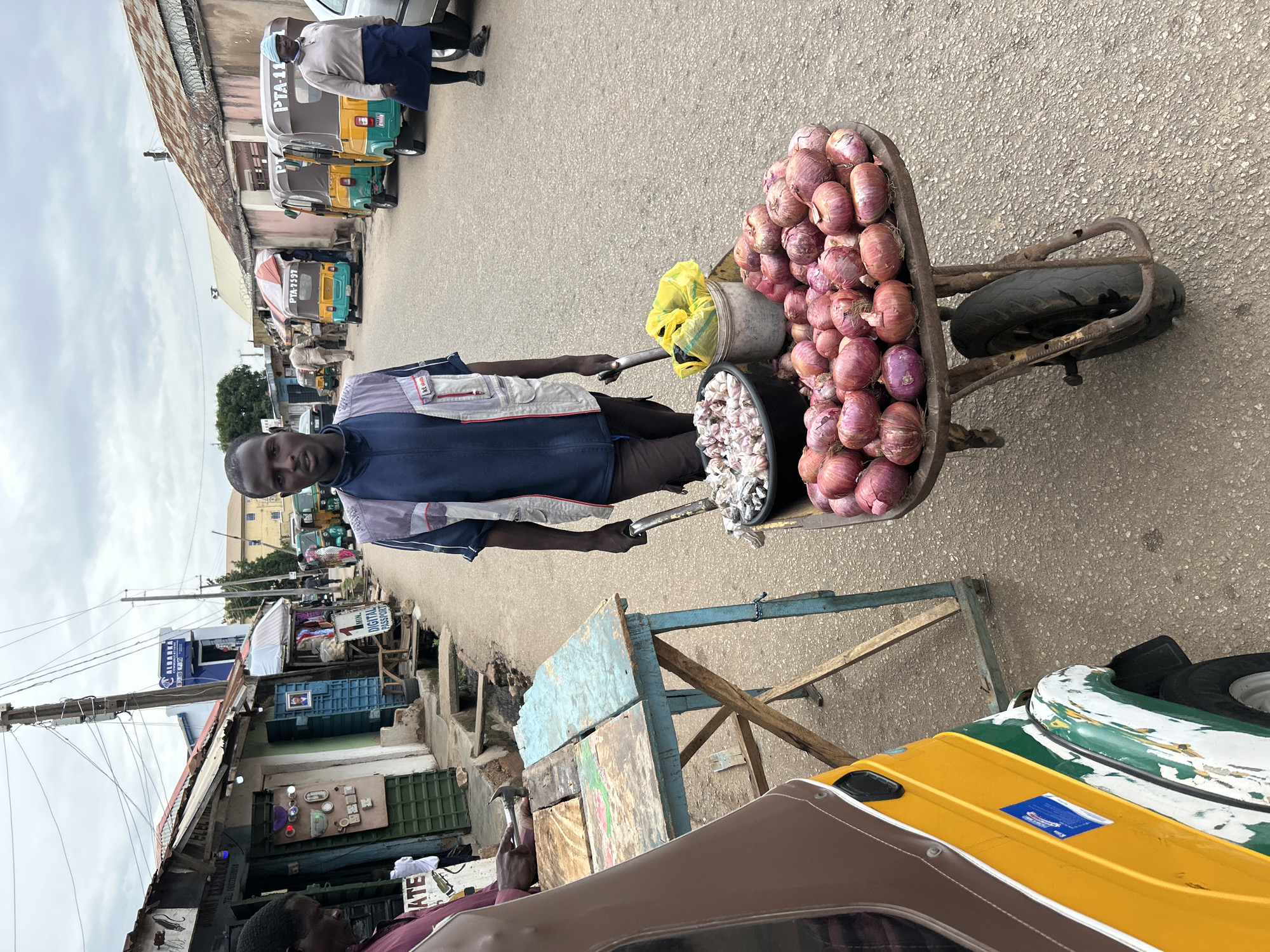A man stands on a paved road, pushing a wheelbarrow filled with large onions and a bucket of garlic. He is wearing a blue and white patterned shirt and dark pants. The wheelbarrow is rusty and has one wheel. Behind him, other people are visible, as well as several motorized rickshaws parked along the side of the road. The scene appears to be a market or a busy street in a town or city. Buildings with corrugated metal roofs line the street, and some of the buildings have signs. One sign has the text "PTA-11" and another has "PTA-1597" visible, suggesting vehicle registration numbers. There is also a sign that reads "MINI DIGITAL PASSPORT" and another partial sign "ALBARRA". The sky is overcast, suggesting cloudy weather. The overall impression is of an active street scene in a populated area.