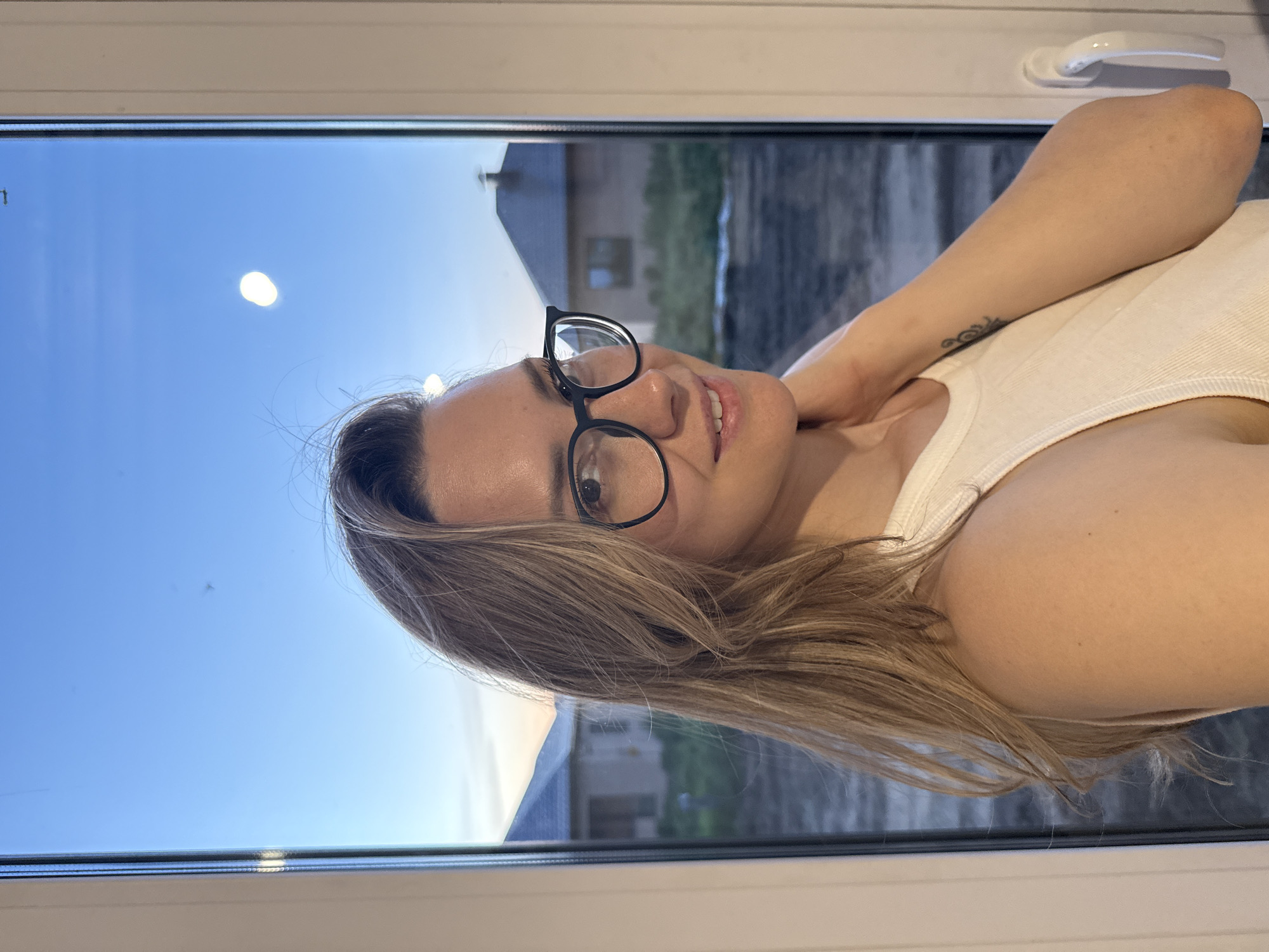 A woman with blonde hair and wearing glasses is visible, posing near a window. Outside the window, buildings are visible, suggesting an urban setting. The location is the city of Chaikovsky, Russia. The woman is wearing a white tank top. She is touching her neck with her right hand. A tattoo is visible on her forearm.