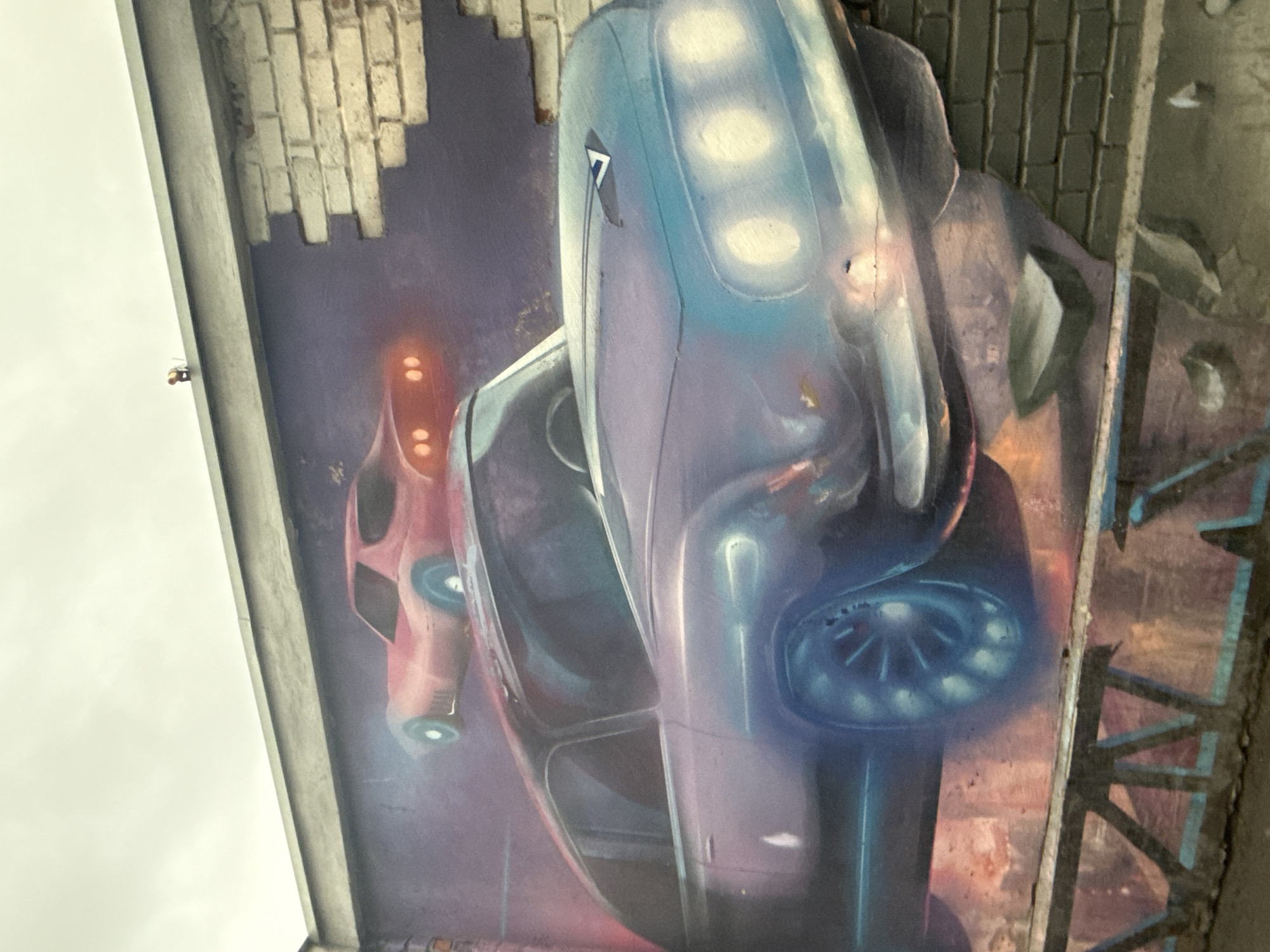 This is a photograph of a section of a mural depicting two stylized vehicles with futuristic designs, possibly robots or spacecraft. The mural is located on the underside of a bridge or similar overpass, as evidenced by the support beams and brickwork surrounding the painted area. The image is oriented at an angle, looking up at the underside of the structure.
The larger vehicle on the right is colored primarily in shades of blue and purple, with illuminated areas that suggest lights or reflections. The smaller vehicle on the left is primarily pink and red with similar light effects. The background of the mural is a mix of purple and brick-like textures. The condition of the mural shows signs of aging or wear, with cracks and imperfections visible on the painted surface. The photograph shows signs of weather conditions like cloudiness, and sky is partly visible on the left side.
Given the presence of the graffiti and urban environment, the location is most likely an area with public art or street art in Saint Petersburg, Russia.