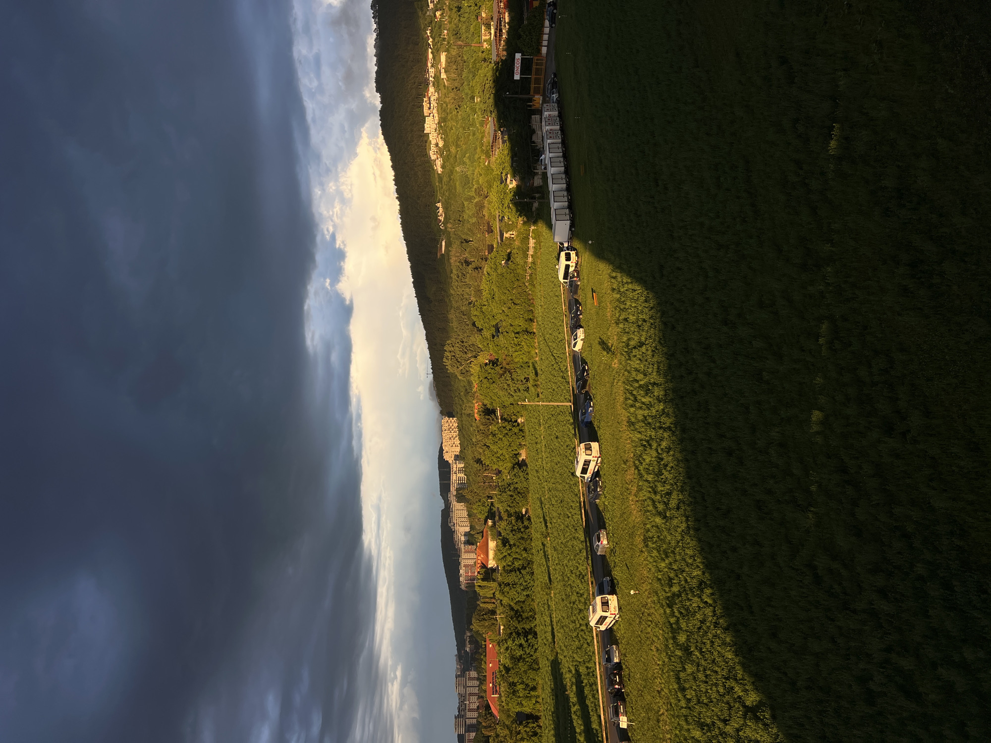 The image, taken from an aerial perspective in Florești, Romania, depicts a landscape at what appears to be either sunrise or sunset, given the strong shadows cast across the grassy field and the golden light illuminating the clouds.
The scene is divided into two distinct halves. On the left, a dramatic sky fills the frame, characterized by dark, ominous storm clouds that gradually transition into lighter, sunlit clouds towards the right.
The right half of the image showcases a long, green grassy field bordered by a road. Several white vehicles, possibly camper vans or recreational vehicles, are parked in a line along the road. A few other cars are also visible. Beyond the road, a hillside is densely covered with trees and dotted with buildings, suggesting a residential area or a town nestled against the natural landscape. The golden hour light highlights the textures of the grass and the contours of the hillside.
The overall atmosphere is one of natural beauty and quietude, with the dynamic sky contrasting with the serene earthly elements. No specific activity or event is clearly identifiable, other than the vehicles being parked.
There is a sign visible on the hillside with the word "ONCOS" written on it.