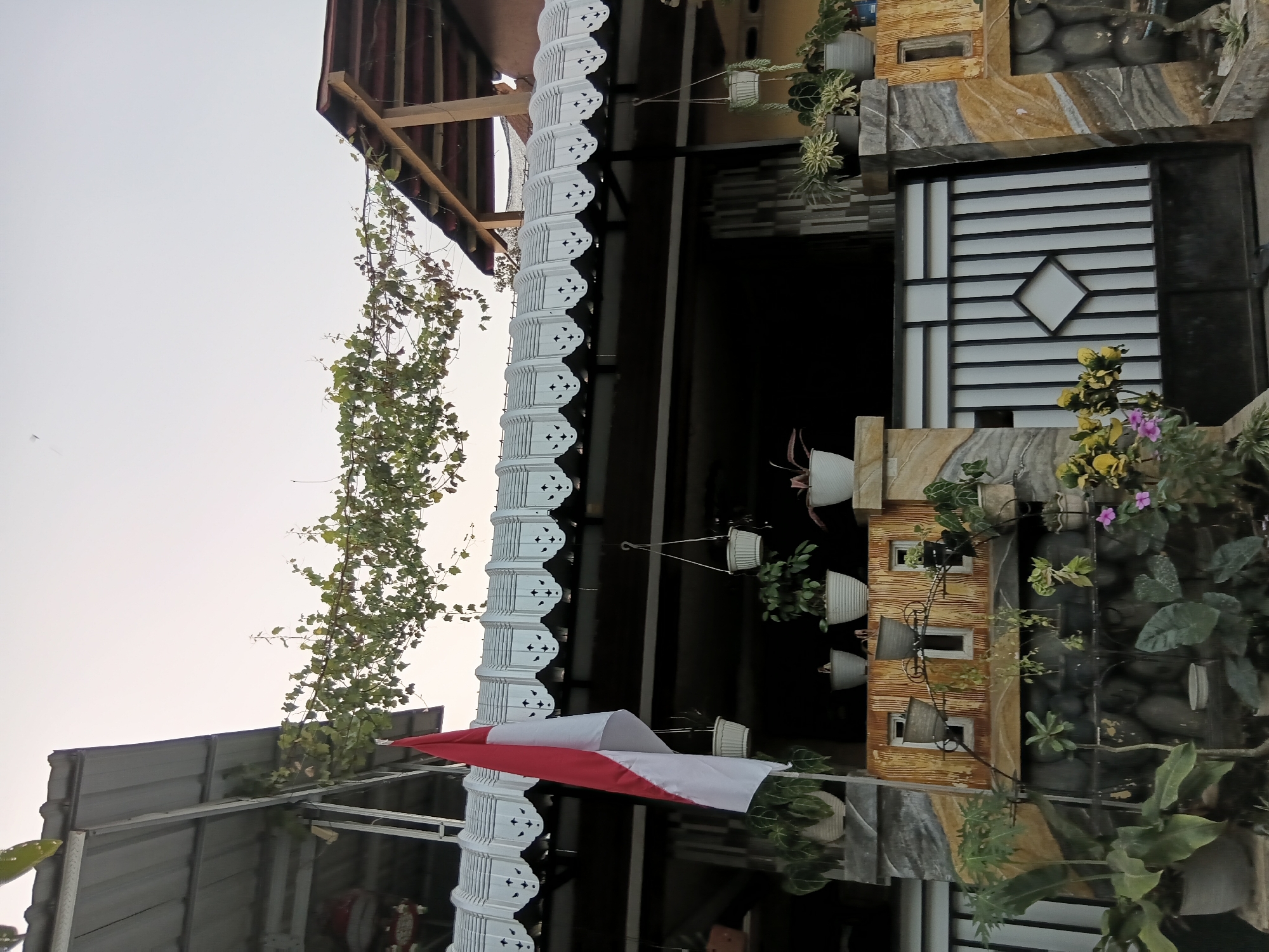 A view of the front of a house in Tanggul Wetan, Indonesia, features a red and white Indonesian flag hanging from a pole. The house has a corrugated metal roof with decorative white eaves and a wooden-framed upper section visible on the right. A vine with green leaves grows across the roofline. Below the roof, a dark opening suggests an entrance or covered area. In front of this, a stone wall with a textured, patterned finish supports a decorative shelf holding several potted plants. A black metal gate with vertical bars and a diamond-shaped cutout is visible in the foreground. Various plants in pots and the ground are scattered throughout the scene, with some small pink and yellow flowers adding color. The overall impression is of a residential property, likely an entryway or facade, decorated with plants and national symbolism. The sky above is overcast.