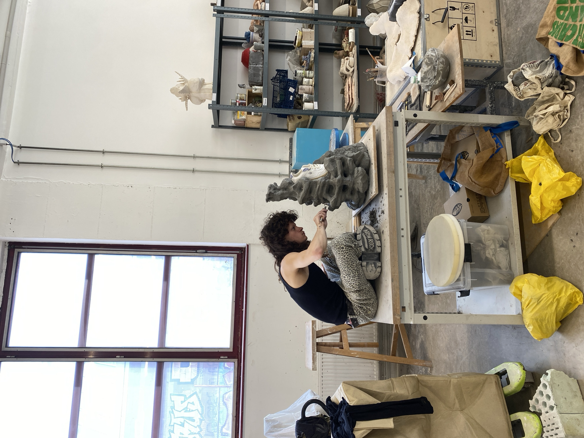 The image captures a vibrant artist's studio or workshop, illuminated by bright natural light streaming from a large window on the left wall, suggesting it is daytime. The primary subject is a young adult with curly dark hair, wearing a black tank top and distinctive patterned trousers, intensely focused on sculpting. They are seated on a wooden stool, meticulously working on a substantial, textured dark grey sculpture, which resembles organic rock formations or abstract masses, placed on a sturdy wooden workbench. The artist uses a small tool to refine the details of the piece. A white athletic shoe rests casually on the grey sculpture, and the artist's own shoe clearly displays "NEW ROCK" on its heel.
The studio environment is functional and shows signs of active work. Against the far wall, a tall metal shelving unit is packed with various art supplies, containers, and smaller sculptural pieces, including a pale, bust-like form on an upper shelf and a blue crate with "MARIA" faintly written on it. Metal pipes run along the upper part of the wall.
The concrete floor is scattered with yellow plastic bags, burlap sacks—one of which partially reads "ORGANI GARDEN"—and other fabric bags. Cardboard boxes, foam fragments, and a black handbag are also visible on the floor and tucked under the tables. Another workbench to the right holds additional materials, including a clear plastic container with a white, circular, cushion-like object. Shipping icons are visible on a wooden crate. The scene conveys a busy, creative process in an artistic workspace, located in Saint-Ouen-sur-Seine, France.