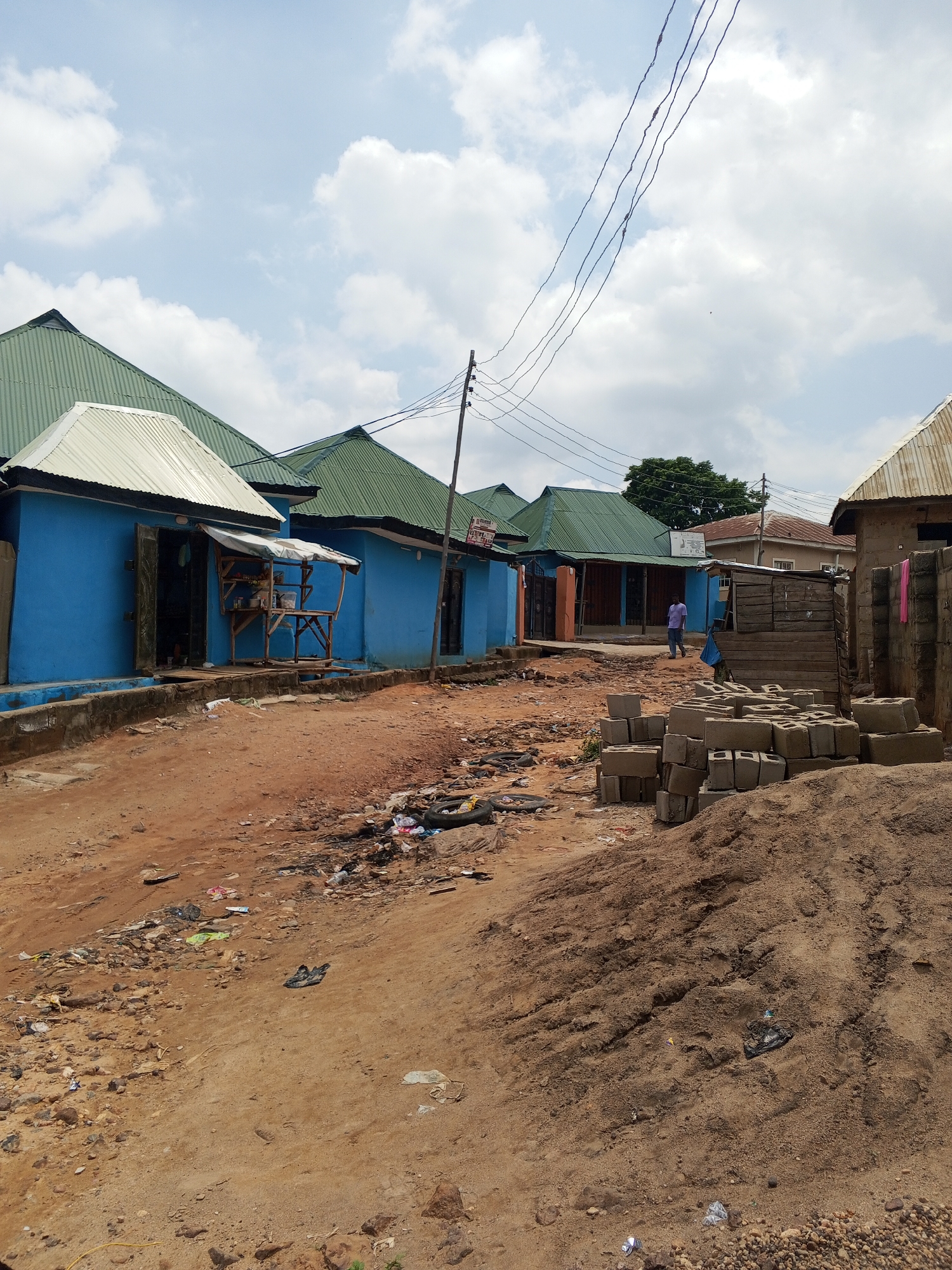 The image depicts a street scene in Lokoja, Nigeria. The street is unpaved, covered in reddish-brown dirt and scattered with trash. There are several one-story buildings along the street, primarily blue with green or white corrugated metal roofs. Some buildings have makeshift stalls or counters in front. A man wearing a light purple shirt and blue jeans is walking down the street in the distance. Piles of cinder blocks and sand are stacked on the right side of the image. Overhead, power lines stretch across the sky, which is partly cloudy. The overall impression is a modest, possibly impoverished neighborhood.