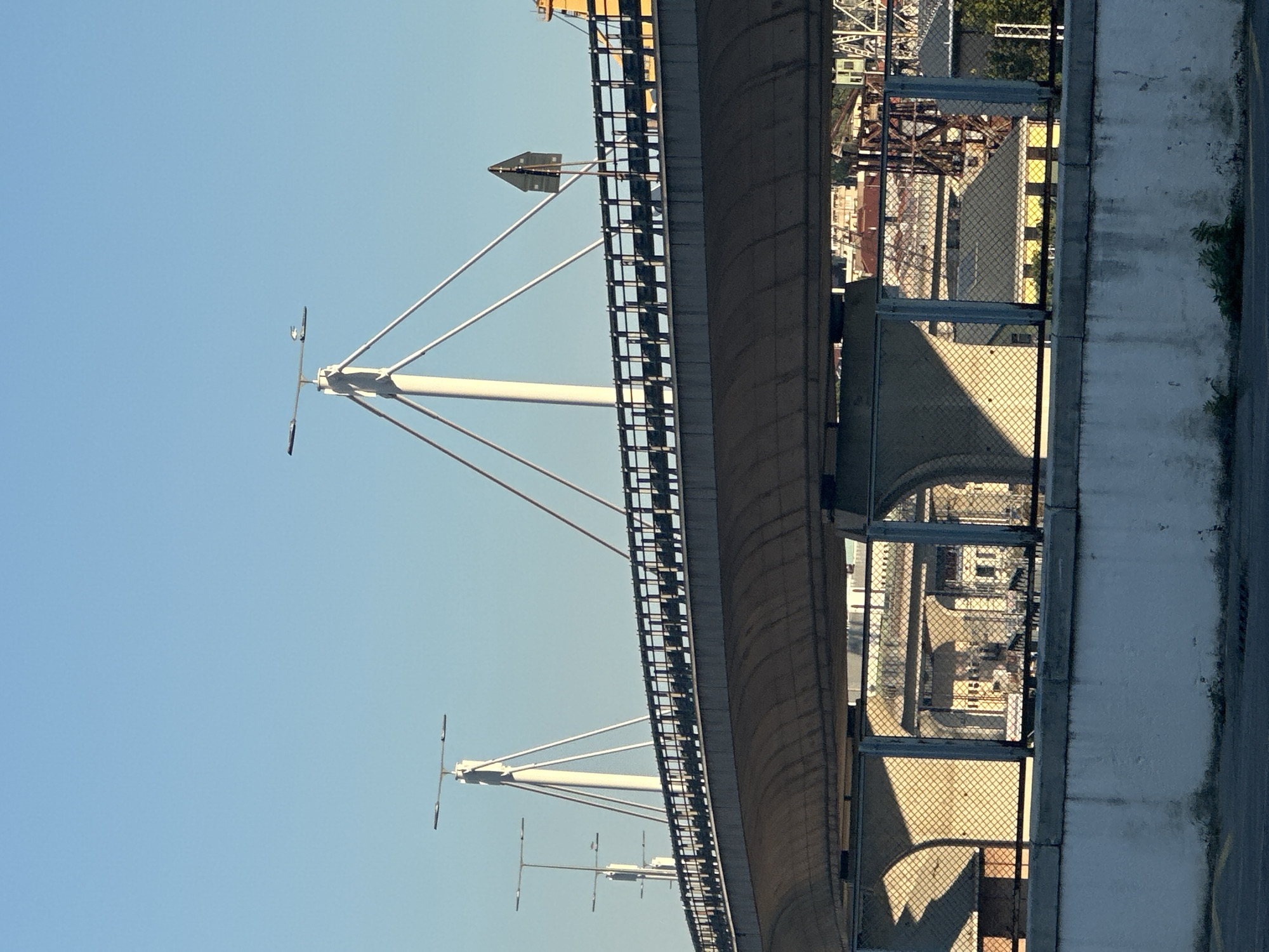 A concrete overpass bridge with a black railing spans across the frame, its underside casting a shadow. The bridge is supported by large concrete pillars and features distinctive white towers with angled cables. On the bridge, a triangular traffic sign is visible. In the background, a clear blue sky dominates, with glimpses of buildings and industrial structures peeking through the bridge's supporting arches, suggesting an urban environment. The scene appears to be in Trieste, Italy, a port city known for its bridges and industrial areas.
