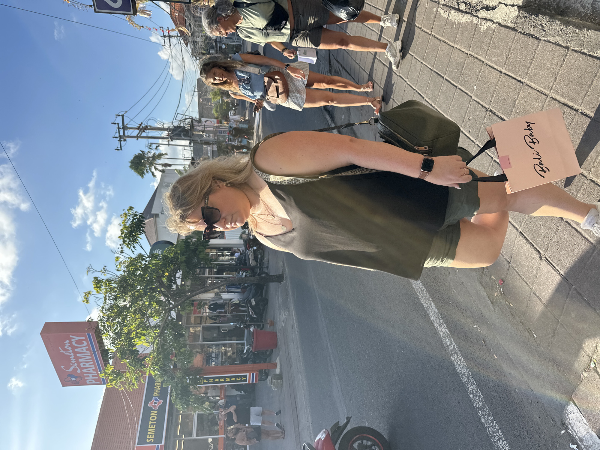 The image captures a street scene in North Kuta, Indonesia, under a clear blue sky with scattered clouds, suggesting it's daytime. The perspective is from ground level, looking upwards and slightly to the right.
In the foreground, a woman with blonde hair is partially visible, wearing sunglasses and a dark top, with a glimpse of her arm and shoulder. She appears to be walking or standing on a paved sidewalk. To her right, a light pink shopping bag with "Bali Baby" written in cursive script is held by its handles. Next to the bag is a dark green backpack. Her hand, wearing a ring and a watch, is also visible.
Behind her, two other women are walking on the sidewalk. The woman in the middle has blonde hair and is wearing a light blue top and a white skirt, carrying a brown bag. The woman further back has gray hair and is wearing a green t-shirt and shorts. They are walking on a brick-patterned sidewalk.
The background shows a street with several shops and businesses. A prominent sign for "SEMETON PHARMACY" is visible, indicating a commercial area. Motorbikes are parked along the side of the street. The scene suggests a typical day in a bustling tourist or shopping district, with people going about their activities. The sunlight indicates it's likely morning or afternoon.