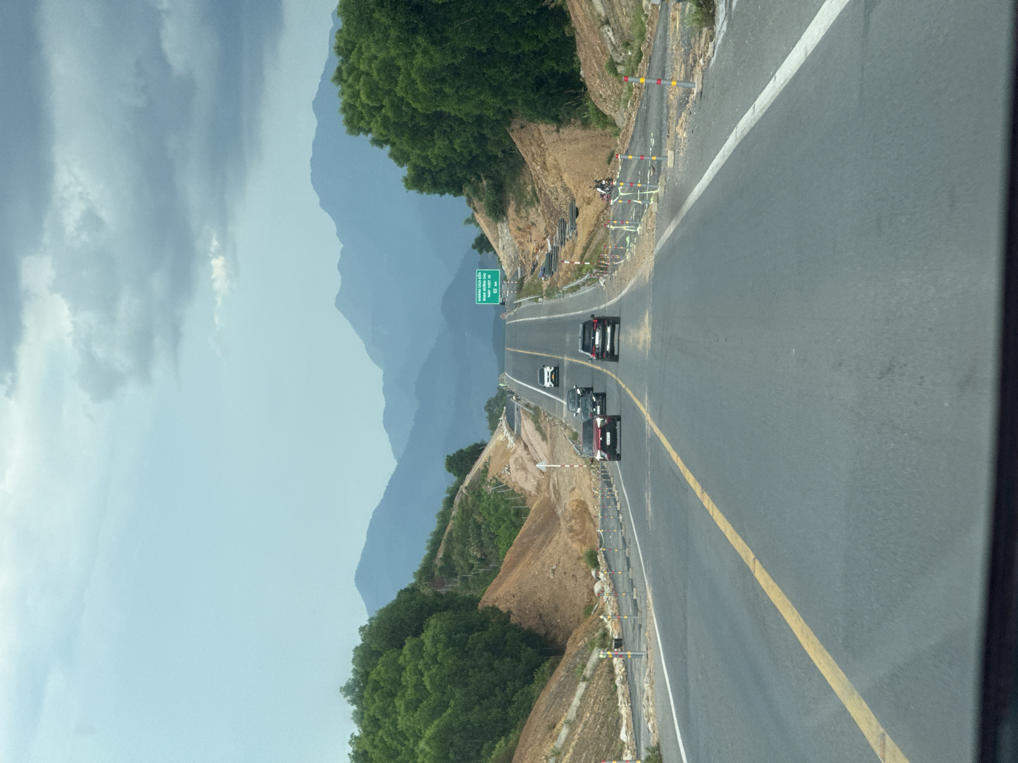 The image shows a highway with several cars driving on it. The highway is surrounded by lush green trees and rolling hills. In the background, mountains can be seen under a cloudy sky. There are also construction barriers and signs along the roadside, indicating that the highway might be under construction or undergoing maintenance. The overall scene suggests a scenic drive through a rural or mountainous area in Vietnam, possibly near Phú Bài Ward.