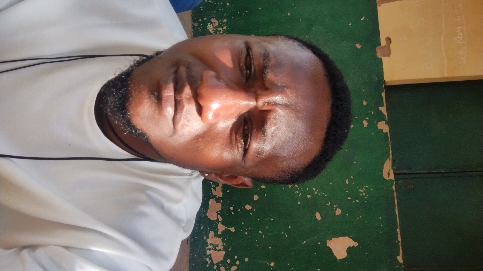 The media file is a close-up, rotated portrait of a Black man. He is wearing a white shirt with a thin black line detail. He has a short beard and dark hair. His skin appears shiny, suggesting it might be oily or sweaty, and he is looking upwards and slightly to the left. The lighting suggests it is daytime, with sunlight casting shadows on his face.
The background is divided into two sections. The left side is a rough, green surface with light brown splatters. The right side is a lighter, yellowish-brown wall with faint markings, possibly writing or drawings. A vertical dark line runs down the middle of this right section, suggesting a seam or divider. Some of the wall's surface is chipped and peeling, revealing a darker layer underneath. The overall scene appears to be outdoors or in a space with a rustic or weathered aesthetic. The context provided places the scene in Jos, Nigeria.
