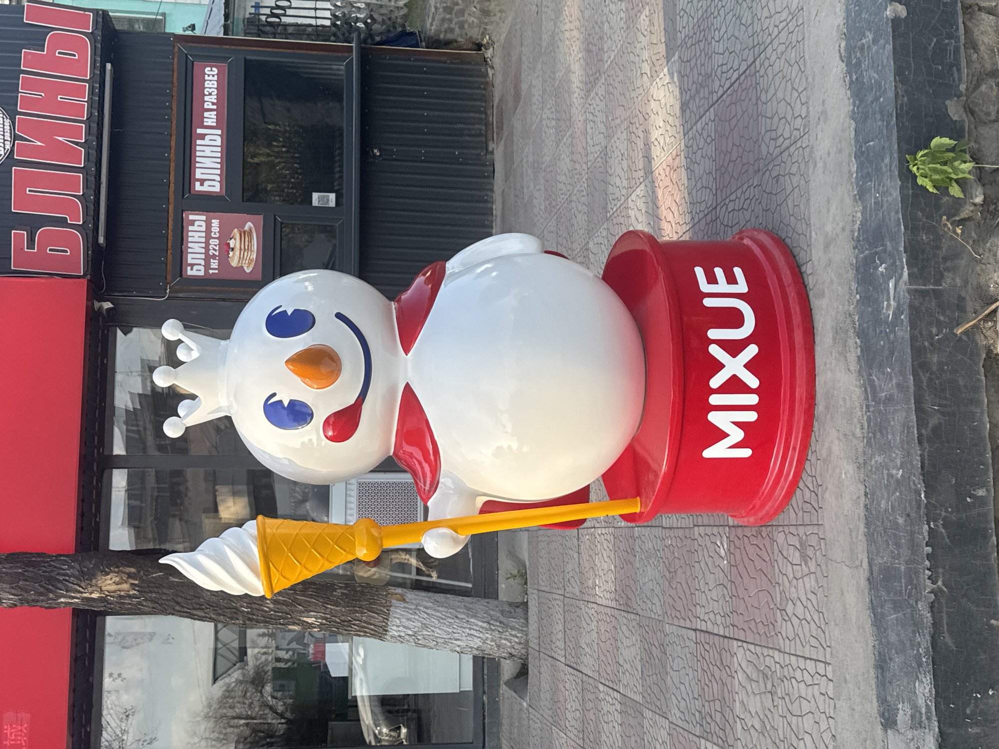 A full-body shot of a white snowman mascot with a red cape and a crown stands on a red circular pedestal with the word "MIXXUE" in white letters. The snowman is holding a yellow ice cream cone with white soft-serve ice cream. Behind the snowman is a building with a red awning and windows. The building has signs in Cyrillic text, indicating it is a store. The ground in front of the store is paved with grey and red patterned tiles. The image was taken in Bishkek, Kyrgyzstan.