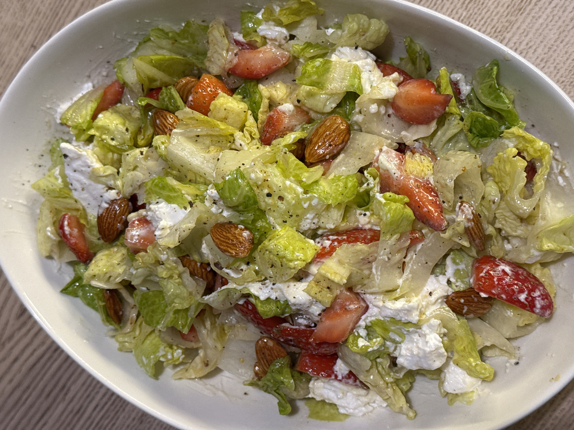 A close-up shot shows a white bowl filled with a salad composed of lettuce, sliced strawberries, almonds, and soft white cheese. The salad is dressed with a light dressing and sprinkled with black pepper. The bowl is placed on a wood-grain surface. The image does not show any identifying information that would associate it with Tel-Aviv, Israel. There are no people, actions, or interactions visible.