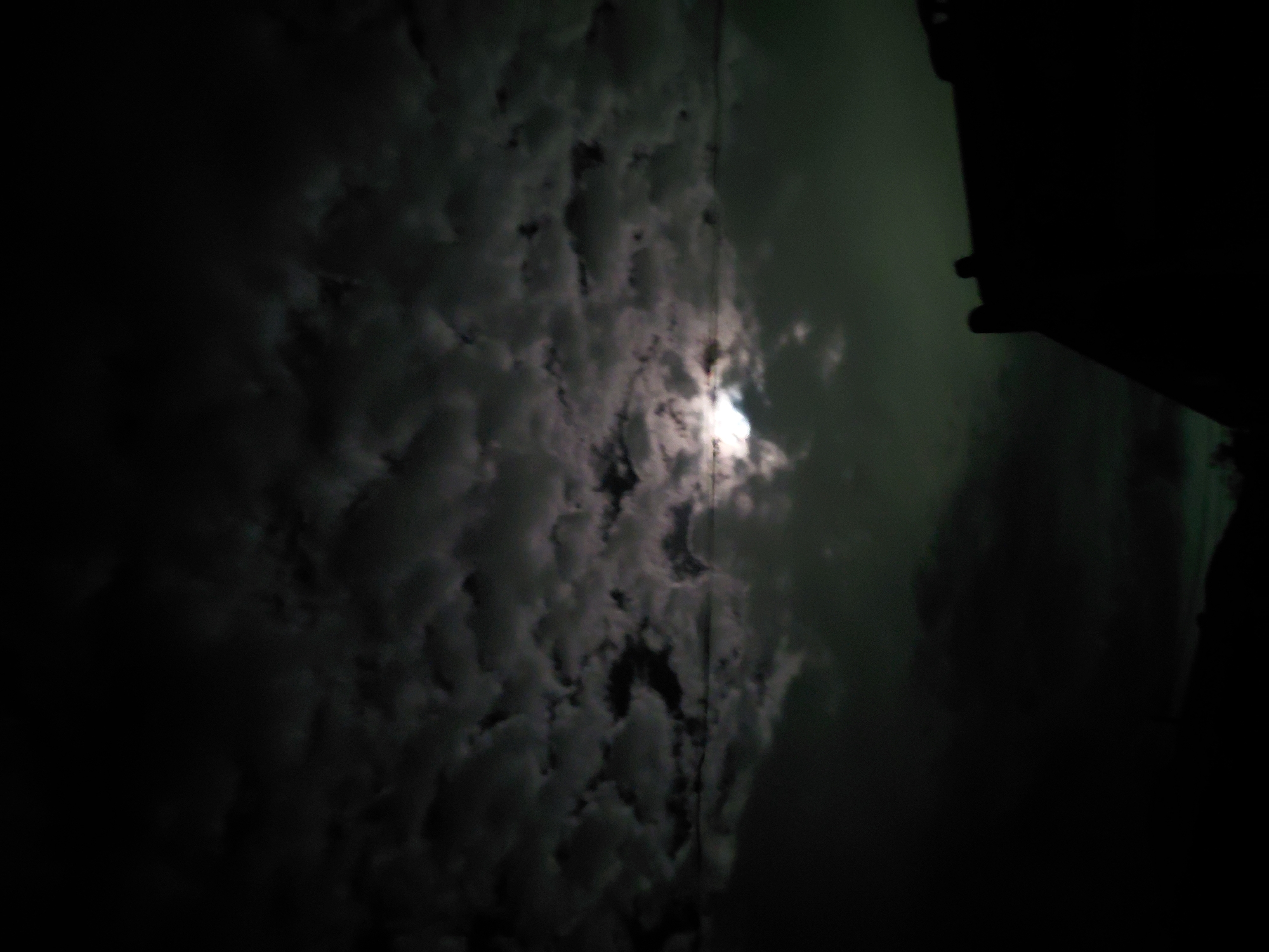 The image displays a nocturnal scene in Santa Ana, Venezuela, dominated by a heavily clouded sky. Dense, dark and grey clouds fill most of the frame, creating a textured canopy. A prominent, bright light source, likely the moon, is visible in the upper-middle right section, piercing through or shining behind the clouds. This light illuminates the surrounding clouds with a soft glow, highlighting their shapes and variations in density.
On the far right side of the image, a stark, dark silhouette of an unidentified structure or object is visible, extending from the top edge downwards. Its form is indistinct due to the extreme darkness, but it appears to be a vertical element like a building edge, pole, or architectural feature. Faint vertical lines, possibly utility wires or cables, can be discerned running through the cloudscape in some areas.
The time of day is clearly night, characterized by the dark sky and the single, strong light source. The weather is cloudy. No people, discernible activities, or visible text are present in the image.