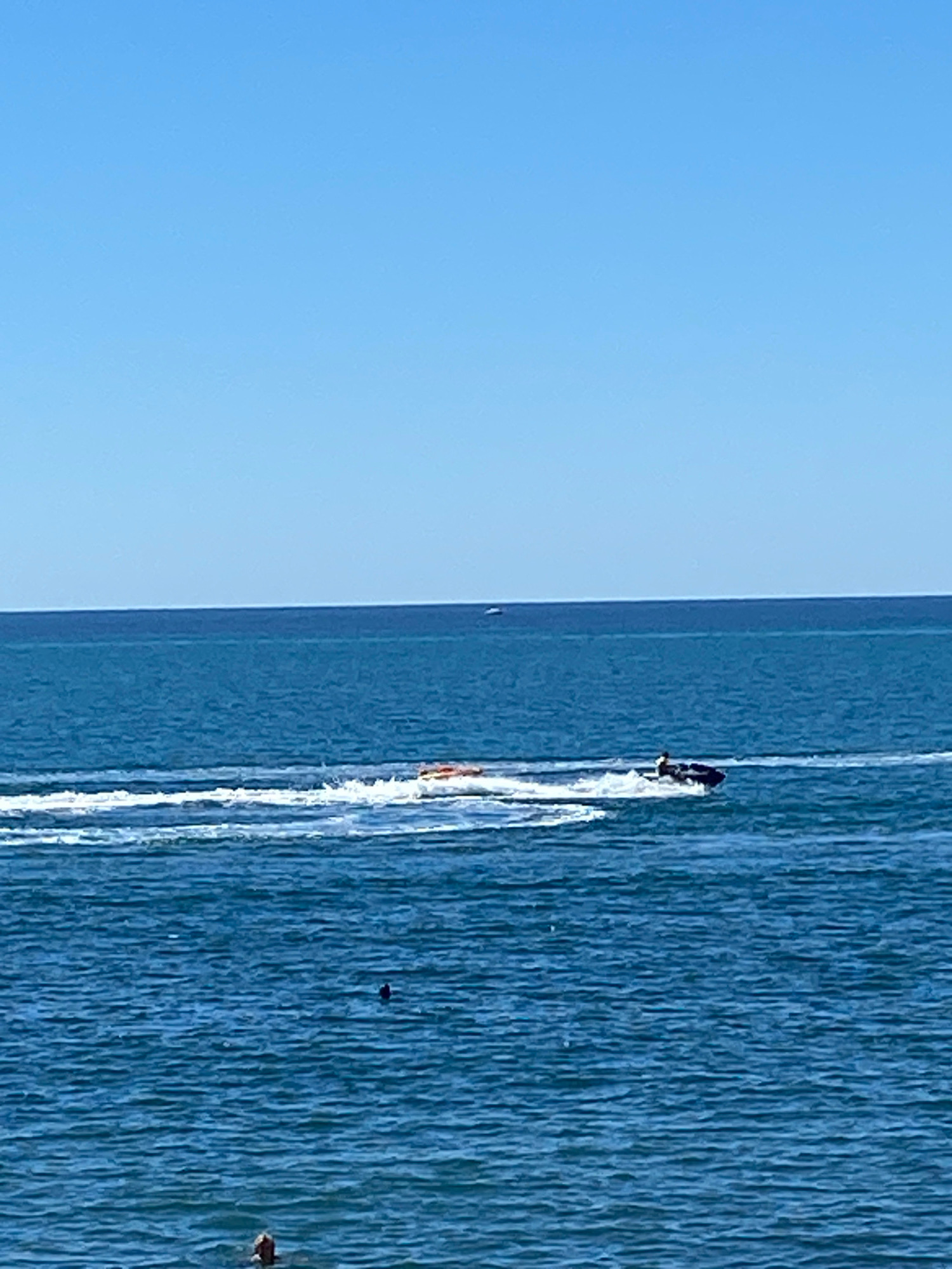 A wide shot captures a bright, clear day at sea, likely the Black Sea off the coast of Sochi, Russia. The sky is a vibrant, cloudless blue, meeting a calm, deep blue ocean at a sharp horizon line. In the mid-ground, a jet ski, black and ridden by a person, cuts through the water, leaving a white wake. To the left of the jet ski, an orange inflatable tube is being towed, also creating a frothy trail in the water. In the further distance, a few small, indistinct boats or buoys dot the horizon. Closer to the foreground, a person's head and shoulders are visible in the water, seemingly swimming. Another dark, small object, possibly a swimmer or a buoy, is also present in the water further out. The overall scene depicts a recreational day at sea.