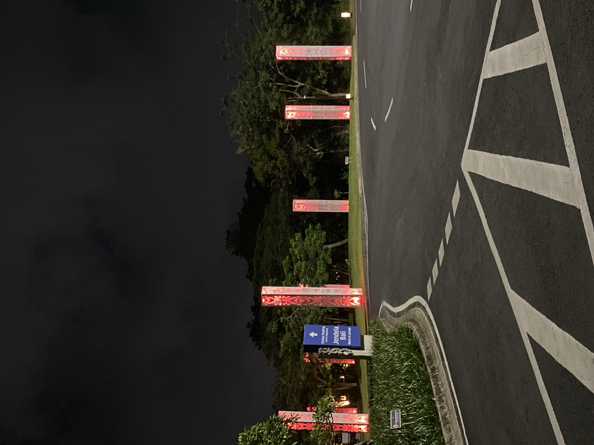 The image depicts an outdoor scene at night, likely an entrance to a resort or hotel. A paved road curves into the foreground, marked with white directional lines. On the left side of the road, a manicured patch of grass is bordered by a concrete curb. A small sign with a symbol and text is planted in the grass.
Further back, a blue directional sign points upwards, indicating "Visitor Parking" and "Jendela Bali Resto & Lounge." Lining the road are several tall, rectangular pillars illuminated from within with a red, intricate pattern. Behind the pillars and the road, lush green trees and foliage form a dense backdrop against a dark, cloudy sky. The scene is lit primarily by the glowing pillars and a few small, ground-level lights. No people are visible in the image. The overall impression is one of a well-maintained, possibly luxurious, nighttime ambiance at a destination named "Jendela Bali."
