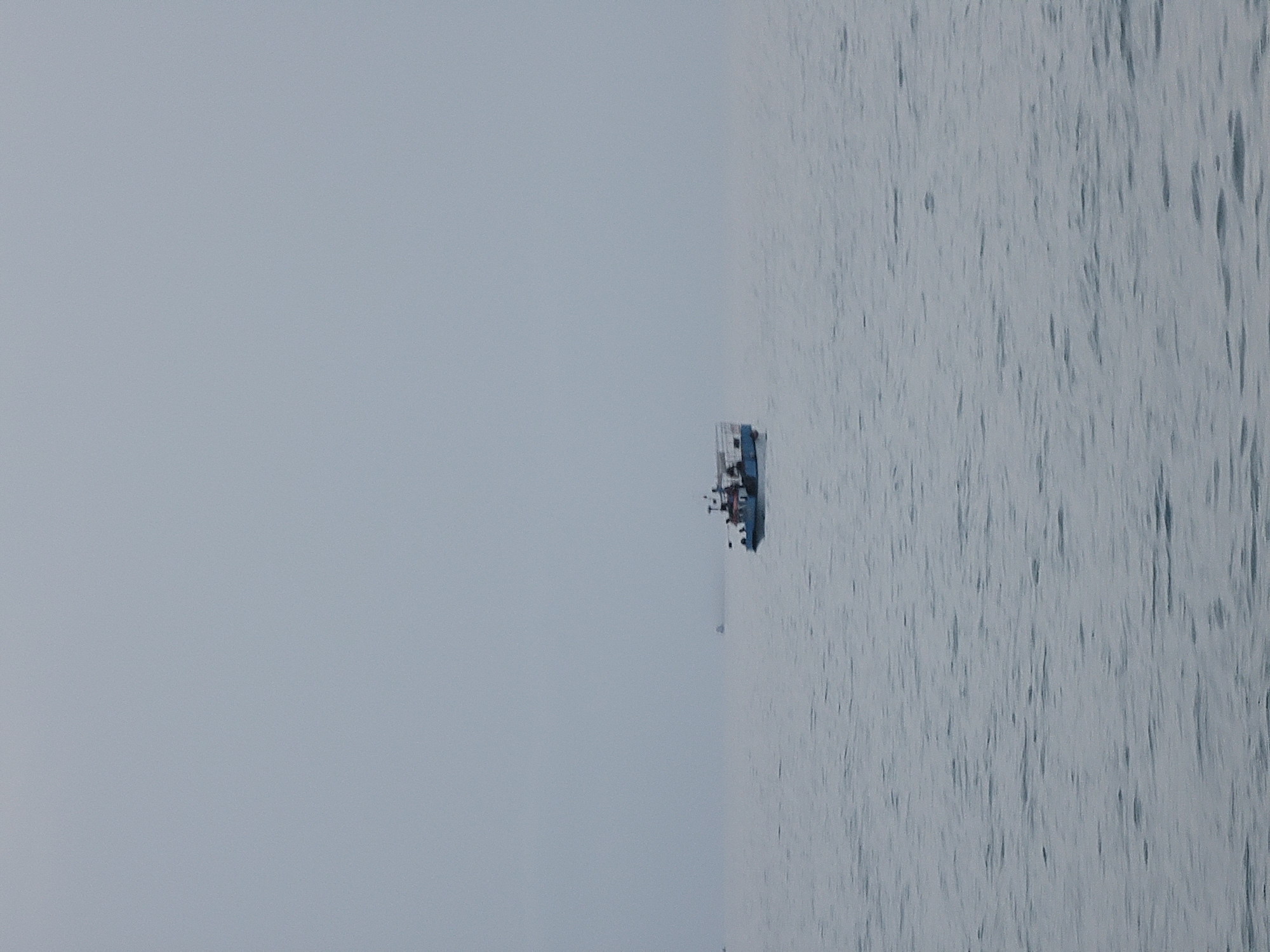 A distant view of a blue and white boat is captured on a calm, grey sea under an overcast sky. The boat, positioned slightly right of center, appears to be a fishing vessel with a cabin structure and a few figures visible on deck. The water is a muted blue-grey with gentle ripples. In the far distance, another faint vessel can be seen. The scene evokes a sense of quietness and perhaps a slightly hazy or foggy atmosphere. The location is the coast of Quarteira, Portugal.