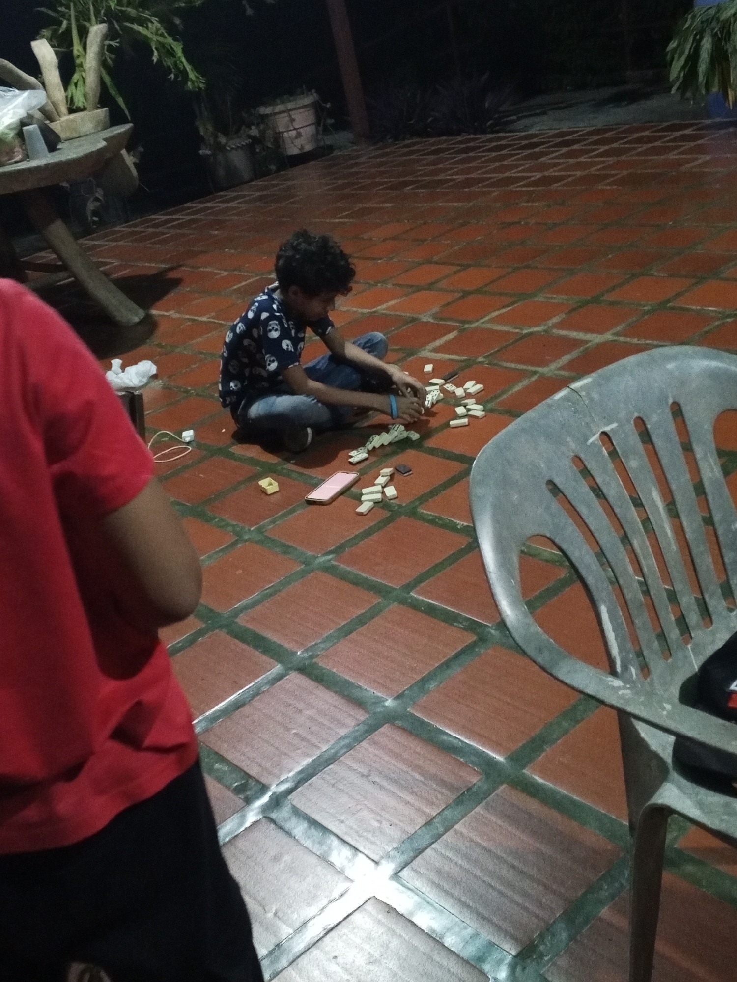 The scene is an outdoor patio in Tinaquillo, Venezuela. The patio is tiled with square, reddish-brown tiles with gray grout. A young boy with curly hair, wearing a blue shirt with skull patterns and jeans, is sitting on the tiles. He is surrounded by dominoes, and it appears he is playing with them. Nearby, on the tile floor, there is a pink smartphone and a pair of wired earphones. To the left, a person in a red shirt and black pants is partially visible. To the right of the boy is a gray plastic chair. In the background, there is a wooden table with plants. The setting appears to be a dimly lit backyard.