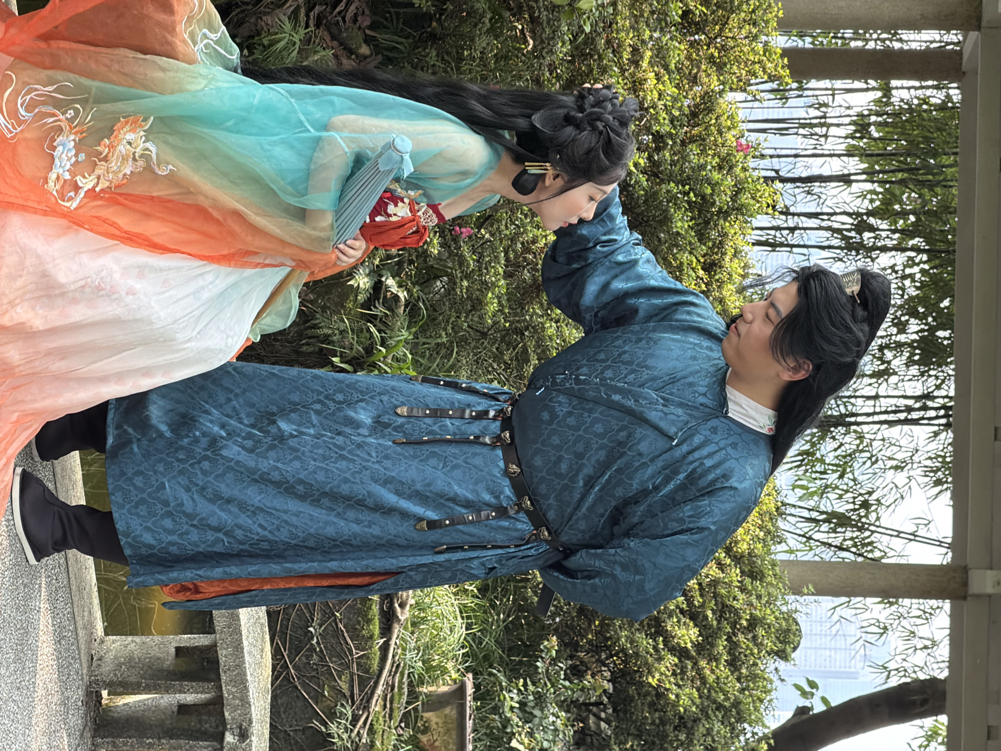 This image depicts two individuals in elaborate traditional Chinese attire engaged in an intimate moment within a garden or park setting. The scene is located in the city of Yuzhong District, China, indicated by the blend of natural greenery and distant urban architecture.
The man, positioned on the right, stands and leans forward, his right hand gently touching the woman's head. He wears a long, dark teal robe with a subtle patterned texture, featuring an ornate black belt with metallic accents and dangling straps. An orange lining is visible through a slit in his robe, and he wears dark footwear. His long black hair is styled into an elevated bun secured with a silver hairpin.
The woman, seated on a grey stone bench on the left, looks up at the man. She is dressed in a multi-layered outfit consisting of a white inner garment and a sheer outer layer that transitions in color from turquoise at the top to orange at the bottom. The orange section is adorned with intricate embroidery depicting mythical creatures, possibly phoenixes. A red fabric element is visible around her torso. Her long black hair is styled with complex braids and buns, and she wears long, black and gold earrings. She holds a closed, light blue pleated fan in her right hand.
The immediate environment includes lush green bushes, some with small pink flowers, and a glimpse of moss-covered roots and a water body near the man's feet. In the background, tall bamboo trees form a screen, and beyond them, modern high-rise buildings of the city skyline are visible under a bright sky. Overarching architectural elements, including a stone pillar and horizontal beam, suggest a pavilion or covered walkway.