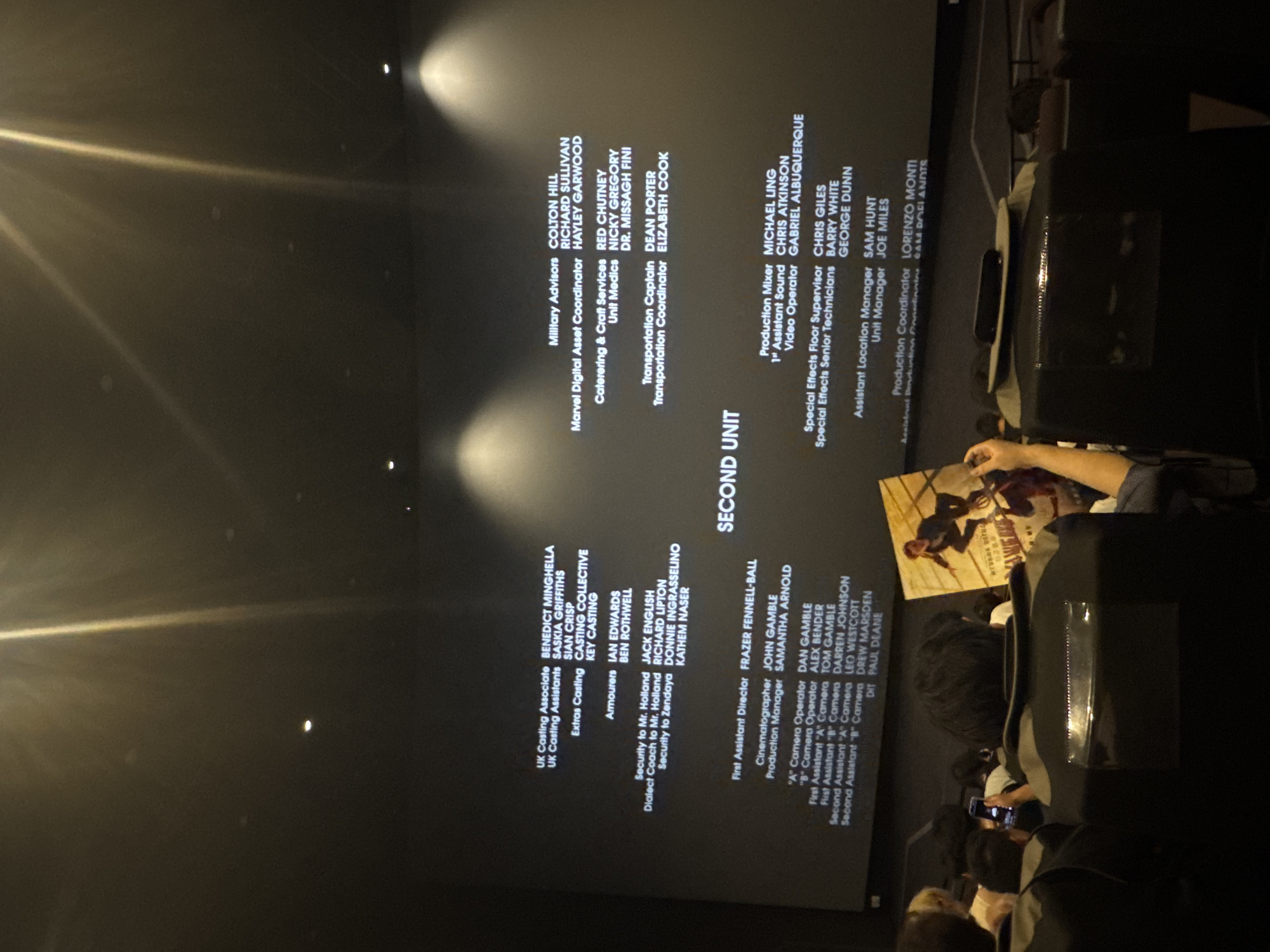 The image shows a movie theater screen with credits rolling, illuminated by stage lights. The credits list various production roles and names. In the foreground, the backs of several people are visible, seated in dark theater chairs. One person on the right is holding up a movie poster. The environment appears to be a dimly lit cinema. The scene is not located in Guta, China, as there is no evidence to support that claim.