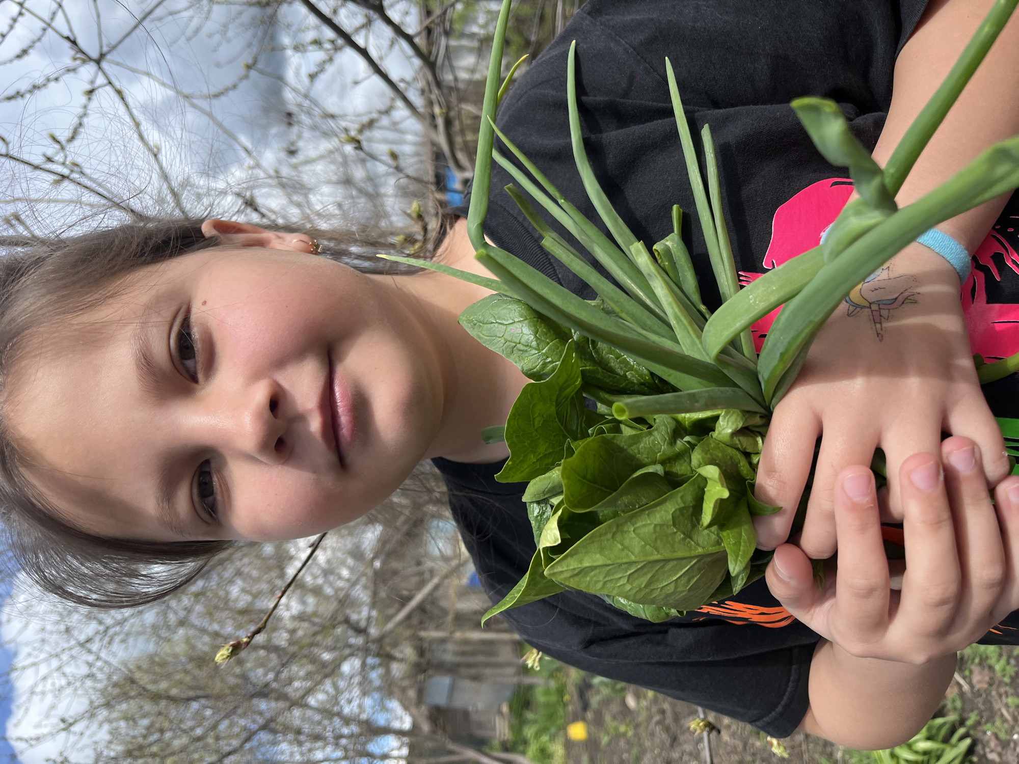 A young girl is holding a bunch of green onions and spinach in her hands. She is wearing a black t-shirt with a design on it. The background shows trees, a cloudy sky, and possibly a garden setting. Based on the prompt, the location is Novopokrovskaya, Russia, but this cannot be confirmed visually from the image itself.