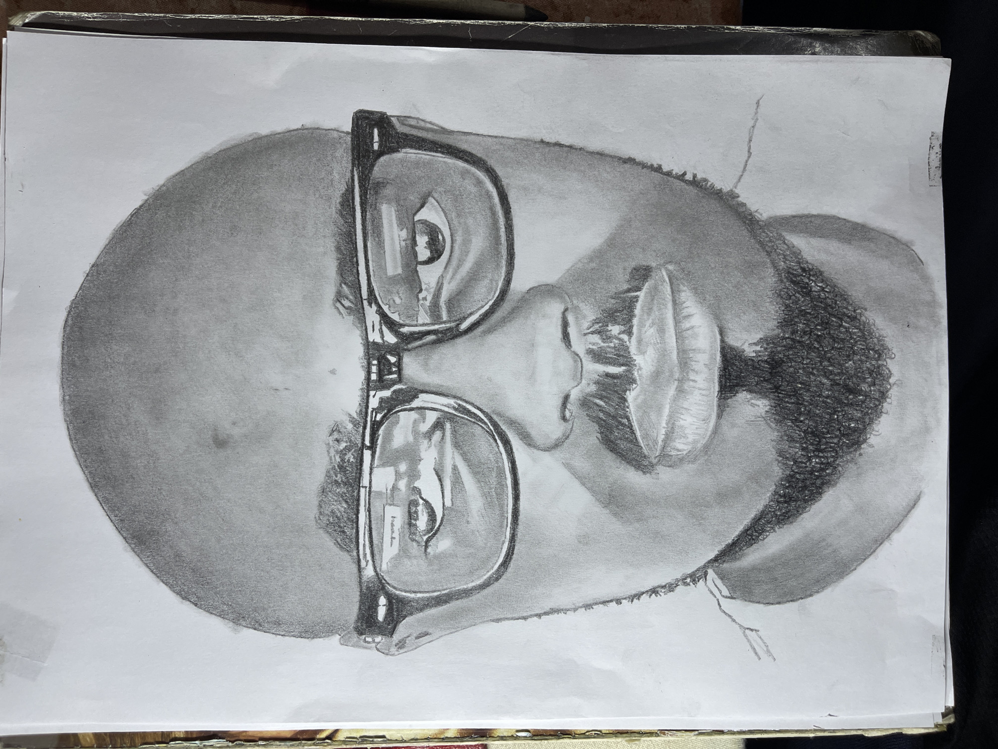The image shows a pencil drawing of a bald man with a beard, wearing glasses. The drawing is on a white sheet of paper. There is no indication in the image that this is located in Jos, Nigeria.