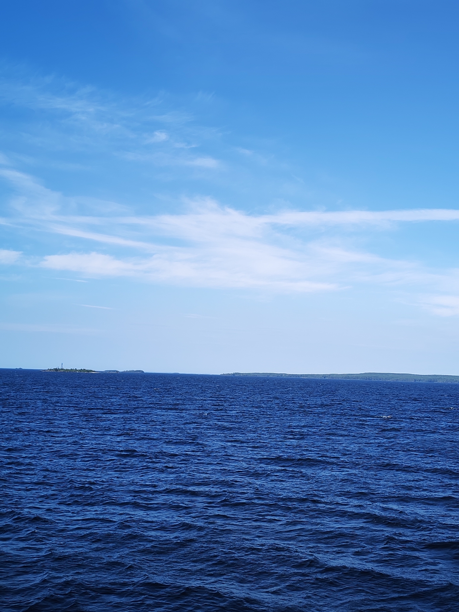 The image depicts a vast expanse of deep blue water, likely a body of water near Shal'skii, Russia. The water is slightly choppy, with ripples suggesting a gentle breeze. In the distance, a long, green landmass is visible along the horizon, hinting at the presence of a coastline or a larger island. On the far left of the horizon, a small island with a tall, white structure, possibly a lighthouse, is situated. The sky above is a clear, bright blue, dotted with wispy white clouds that stretch across the frame. The overall impression is one of a serene and expansive natural landscape.