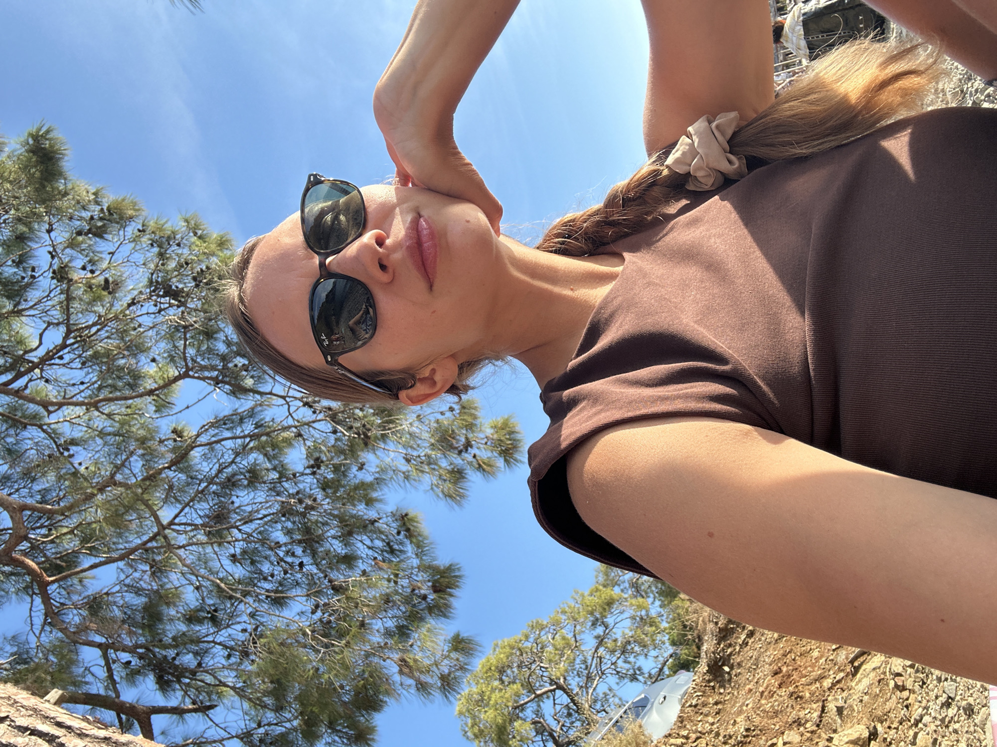 This outdoor selfie, likely taken in Fethiye, Turkey, features a young woman seen from a low angle, looking upwards towards the camera. Her face is centered in the upper mid-frame, with her eyes mostly obscured by dark sunglasses. She has light pink lipstick on her slightly parted lips. Her light brown hair is braided and held back by a beige scrunchie, resting on her shoulder. She is wearing a brown, ribbed short-sleeved top. Her right arm is bent with her hand near her head, creating a posed appearance.
The background is a clear, vibrant blue sky with no clouds, indicating bright, sunny weather. Numerous green pine trees with visible pinecones frame the left and lower portions of the image, suggesting a Mediterranean or forest setting. The ground on the far right is rocky and earthy. Strong, direct sunlight from above casts distinct shadows on her body, especially her arm and shirt, suggesting midday or early afternoon. A partial view of a white object, possibly a vehicle or structure, is visible in the upper right background. The brand "Ray-Ban" is visible on the right lens of her sunglasses.