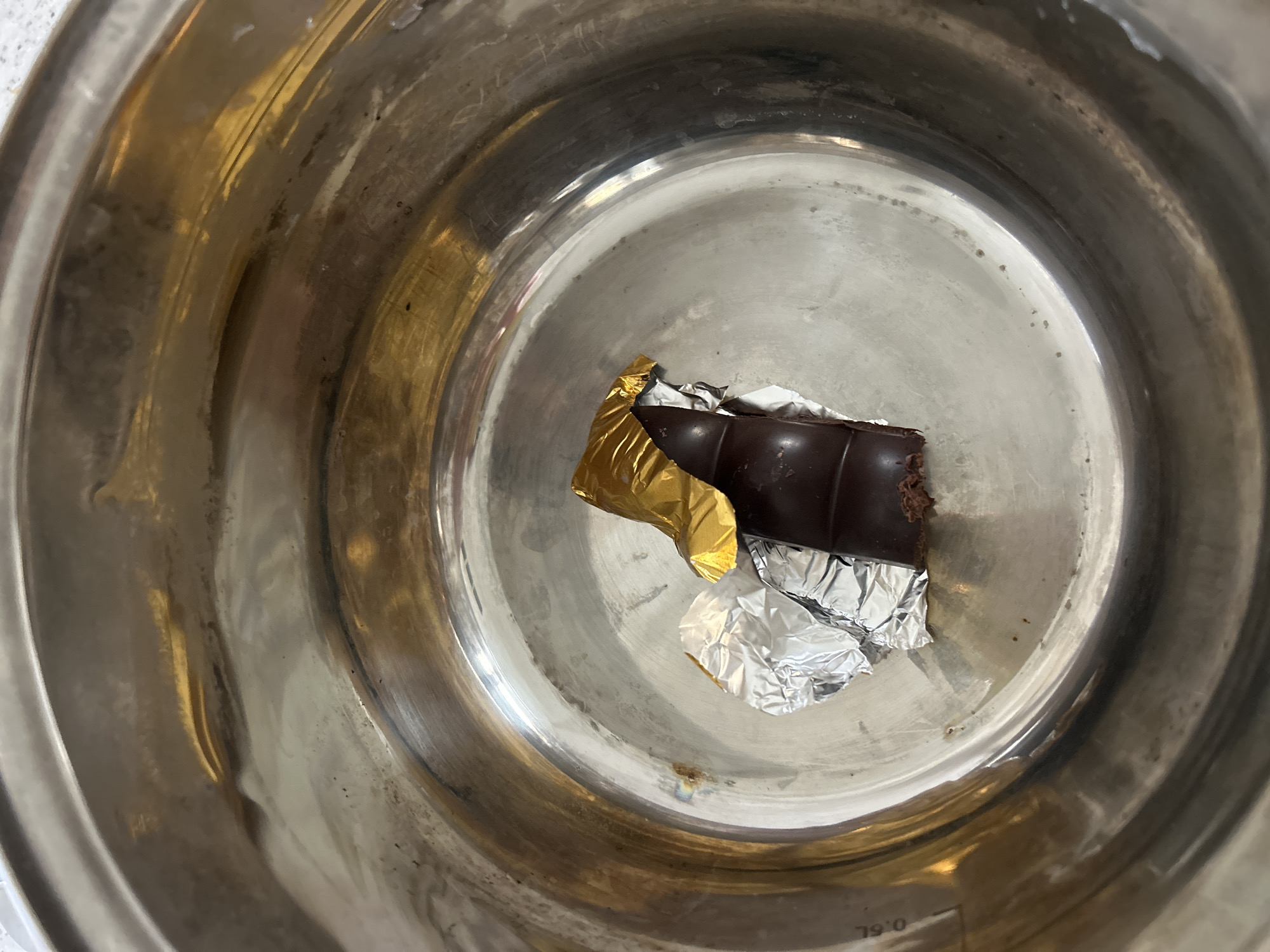 A top-down shot shows a partially unwrapped bar of dark chocolate in a stainless steel pot. The chocolate is wrapped in gold and silver foil. The interior of the pot is clean, with the chocolate positioned in the center.
