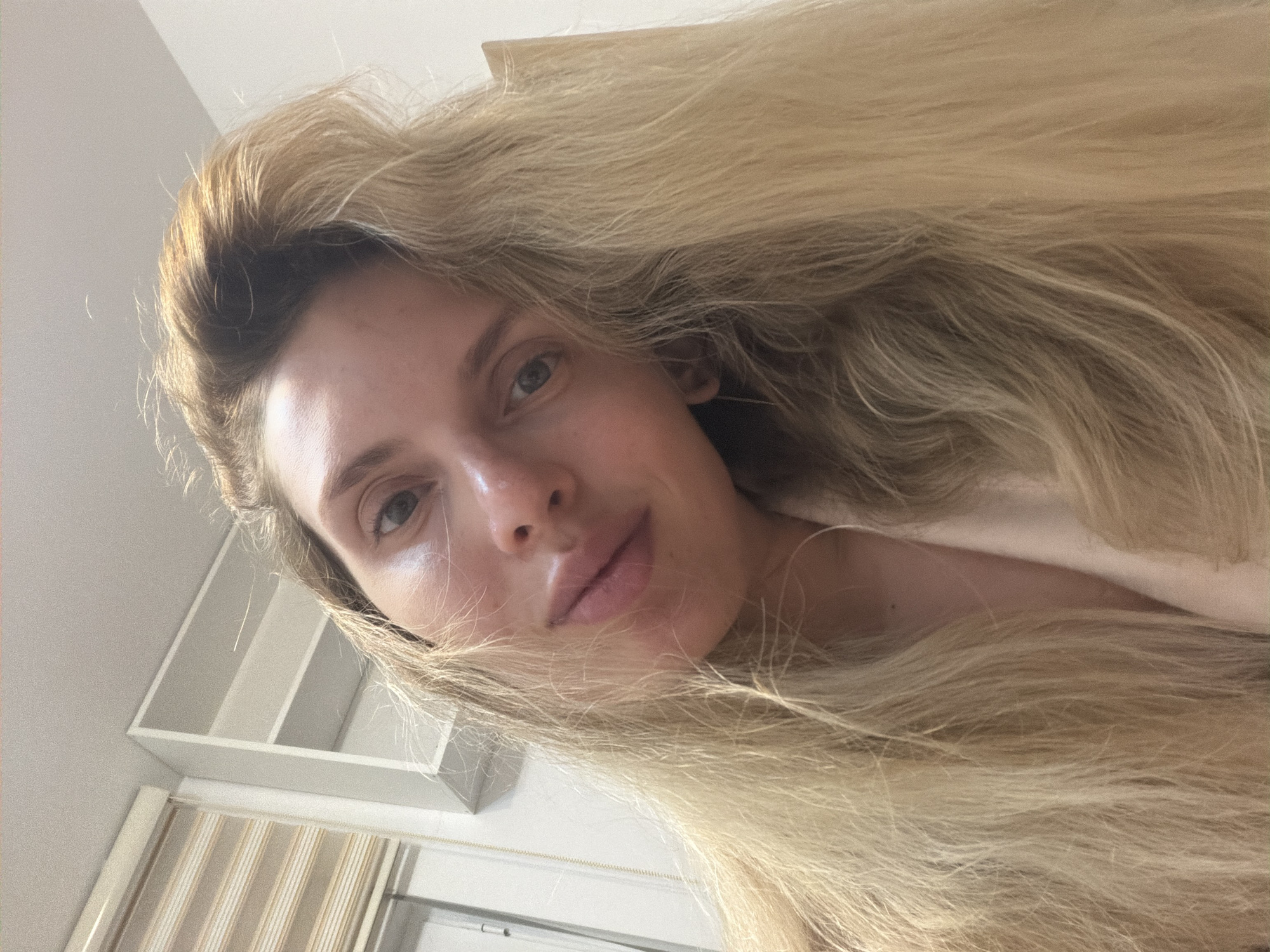 This close-up, low-angle selfie features a young woman with long, voluminous blonde hair that cascades around her face, filling a significant portion of the frame. She has light-colored eyes (appearing blue or green) and full lips, looking directly into the camera with a calm, neutral expression. Her skin shows natural highlights, and she appears to be without makeup. The background, seen from a tilted perspective, includes white or light-toned shelving units or cubbies, and a window with vertical striped blinds or curtains. The setting is indoors, with soft, natural lighting indicating daytime. The photograph captures a moment of stillness, emphasizing the subject's features and hair. Although the image itself does not contain specific geographical markers, it is associated with Budva, Montenegro. There is no visible text.
