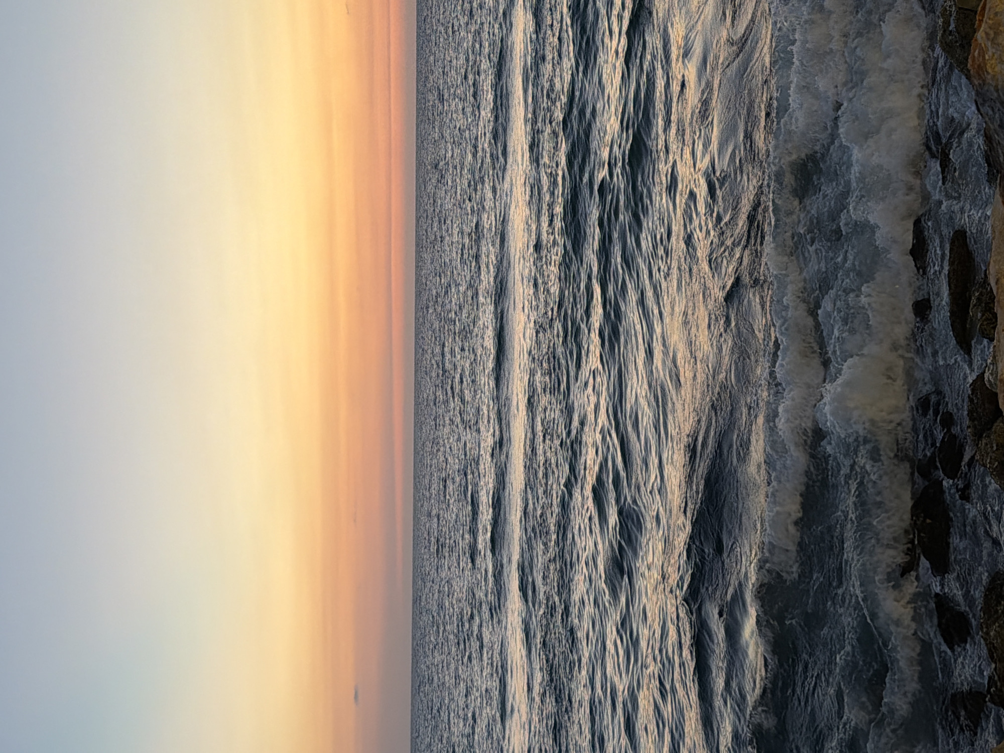The image depicts a view of the Mediterranean Sea during sunset in Tel Aviv, Israel. The left side of the image shows the sky transitioning from light blue to shades of orange and yellow as the sun sets. To the right of the sky, the sea is visible with its surface showing various textures from gentle ripples to more turbulent wave patterns. The far-right edge of the image reveals rocky outcroppings, suggesting a coastal area. There are no people visible in the scene.