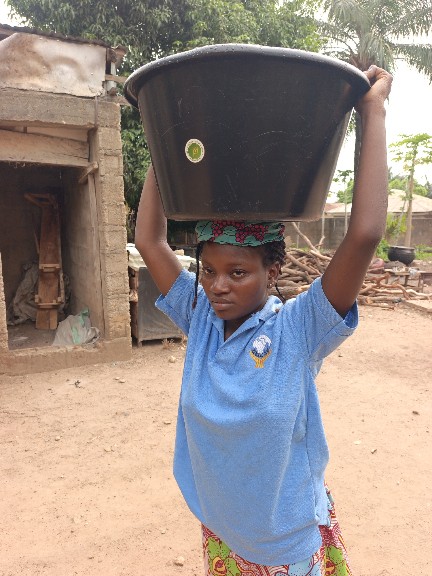 This image depicts a young woman in Akwanga, Nigeria, carrying a large black plastic basin on her head, supporting it with both hands. She wears a light blue polo shirt, a vibrant patterned skirt, and a matching green and red patterned headwrap. Her expression is serious and focused, indicative of the task at hand.
The setting is an outdoor, rural or semi-urban environment with a reddish-brown dirt ground scattered with small stones. In the left background, there is a simple, partially constructed building made of concrete blocks and wooden beams, topped with a corrugated metal roof. Lush green foliage, including palm trees, forms the backdrop, suggesting a tropical climate. To the right, a large pile of cut firewood is visible, alongside other distant structures and a dark pot over what appears to be an outdoor cooking area.
Notable details include a circular emblem on her blue shirt featuring a globe cradled by two hands, with partial text "GLOBAL LEADERSHIP." A circular green and white sticker on the black basin reads "SUSTAINABLE DEVELOPMENT TECHNOLOGY" and "PROMOTING ENERGY EFFICIENCY AND" (partially visible), along with a symbol resembling a recycling or sustainability logo. The lighting indicates daytime, likely under an overcast sky, providing soft, even illumination.