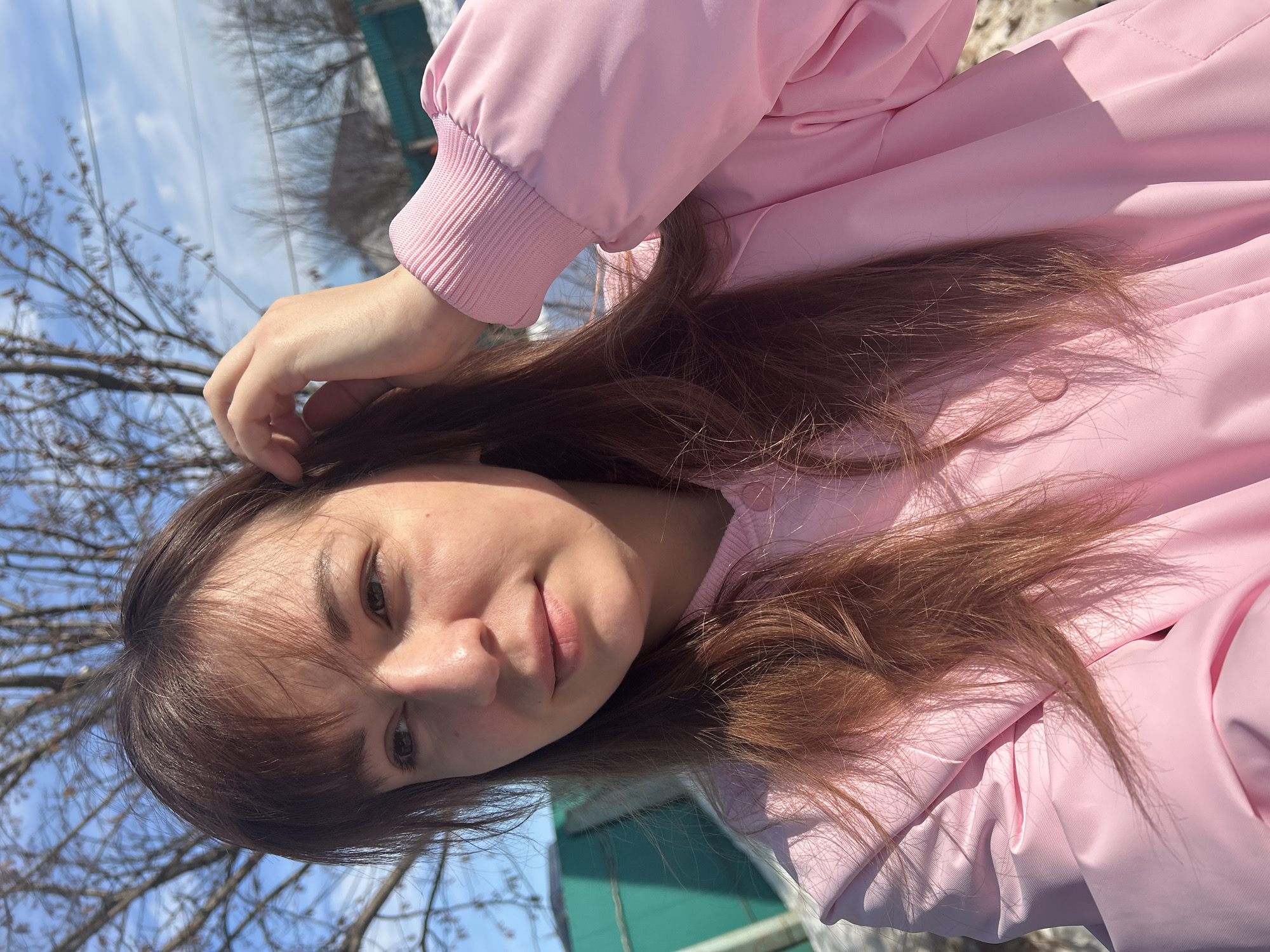 The image displays an adult female, positioned from the chest up and slightly off-center left, outdoors in the city of Фоки, Russia. She has fair skin, dark brown, long hair with bangs, and is wearing a light pink jacket featuring ribbed cuffs on the sleeves. Her right hand is raised, with fingers touching or running through her hair near her right temple. Her gaze is directed slightly to the right of the camera. Strong sunlight illuminates the right side of her face and jacket, casting shadows on her left side and from her hair onto the jacket. The background consists of a clear blue sky with scattered wispy white clouds, bare tree branches, and faint power lines. A dark teal-green fence is visible behind the subject on the left, and patches of snow or ice are present on the ground, indicating a cold season. A building with a white roof is visible in the distant background on the right.