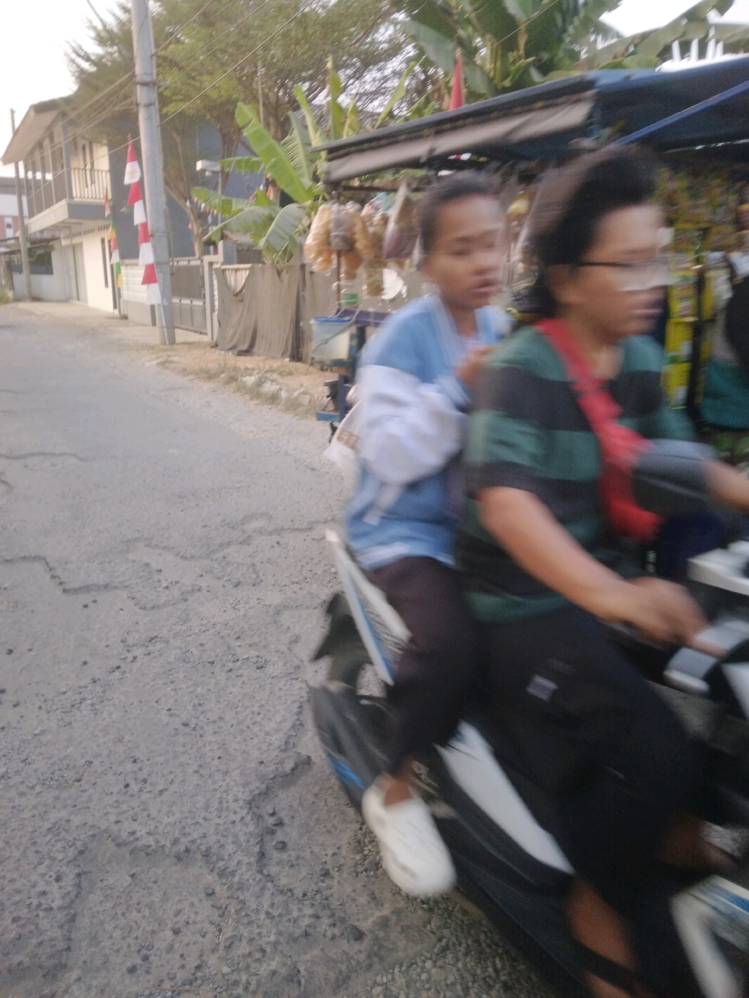 Two individuals are riding on a motorcycle on a paved road. The person in the front is wearing a blue shirt and a white garment over it. The person in the back is wearing a green and black striped shirt with a red strap across their chest and glasses. They are both looking forward. The environment is outdoors with a building and trees visible in the background. The road appears to be in a semi-urban or rural area, indicated by the presence of a building with a balcony, some vegetation including banana trees, and a small roadside stall. The image is slightly blurred, suggesting movement. The flags on the building might indicate a national celebration or local tradition.