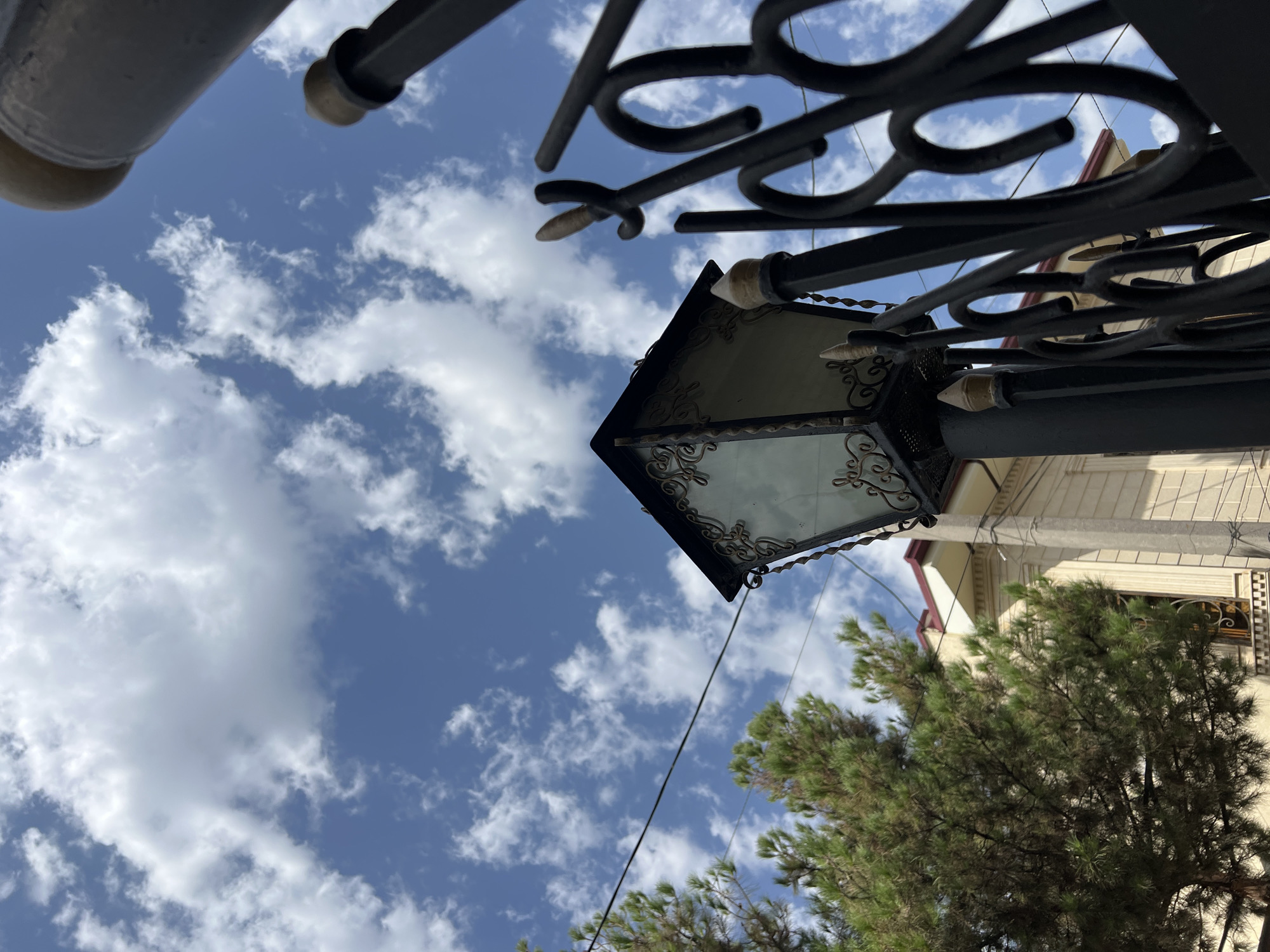 The image depicts a low-angle view looking up at a streetlamp in Tashkent, Uzbekistan. The streetlamp, with its ornate black metalwork and glass panels, stands in the foreground to the left. Behind it, a section of a building with light-colored walls and a red roof is visible. To the left of the streetlamp, a dense green tree is present. A black wrought-iron fence runs along the right side of the frame. The sky above is a clear blue, dotted with scattered white clouds. Power lines are also visible stretching across the sky. The scene suggests an outdoor setting, possibly a residential area or a park in Tashkent, under a daytime sky.