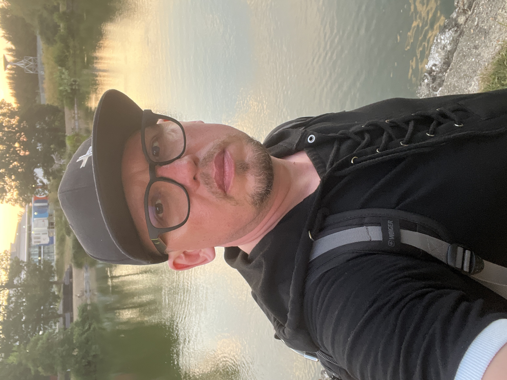 A selfie shows a man with a beard, wearing glasses and a black baseball cap. He is wearing a black hoodie with lace-up detailing and a backpack. The man is standing next to a body of water, possibly a lake or river, with trees and buildings visible in the background. The lighting suggests it is either sunrise or sunset. The location is specified as the River Wey in Guildford, United Kingdom.
