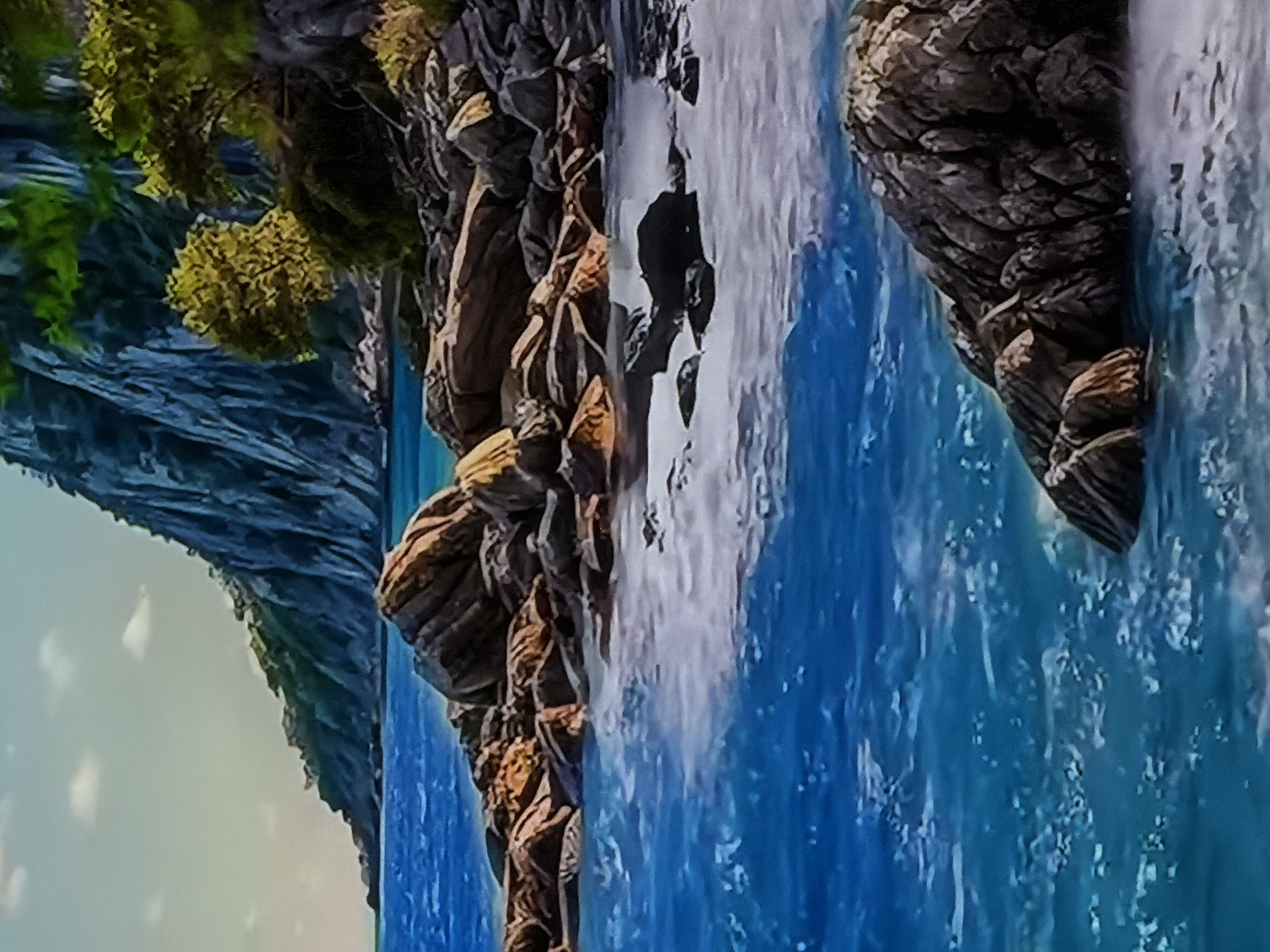 The media file presents a vibrant, digitally rendered or highly stylized natural landscape scene, depicted as if oriented horizontally. The central feature is a majestic waterfall cascading into a bright blue body of water, likely a river or large stream, flanked by rugged terrain.
On the left side of the image, a towering, dark, and intricately textured cliff or mountain rises steeply from the water. Its surface is adorned with patches of lush green and yellowish-green foliage, resembling moss or dense plant growth, particularly at its base and along its contours. The sky above this cliff is a soft, light blue-gray, hinting at a clear or partly cloudy daytime.
The right side of the scene is dominated by a complex arrangement of large, dark, and heavily textured rocks and boulders that form the riverbank. These rock formations display shades of brown, grey, and dark earthy tones, with distinct patterns of stratification and erosion. Green foliage also clings to these rocky outcrops.
A significant waterfall is prominently visible on the right, with bright white, rushing water tumbling vigorously into the blue river below. The river water itself is a striking azure, interspersed with areas of white foam and turbulence, especially where the waterfall impacts the surface, indicating its swift current.
No people, animals, or man-made objects are present in the scene. The landscape appears untouched and wild, conveying a sense of serene natural beauty and grandeur. The illumination suggests it is daytime, likely under a bright sky. There is no visible text. The image's vivid colors and stylized textures contribute to an ethereal and picturesque aesthetic.