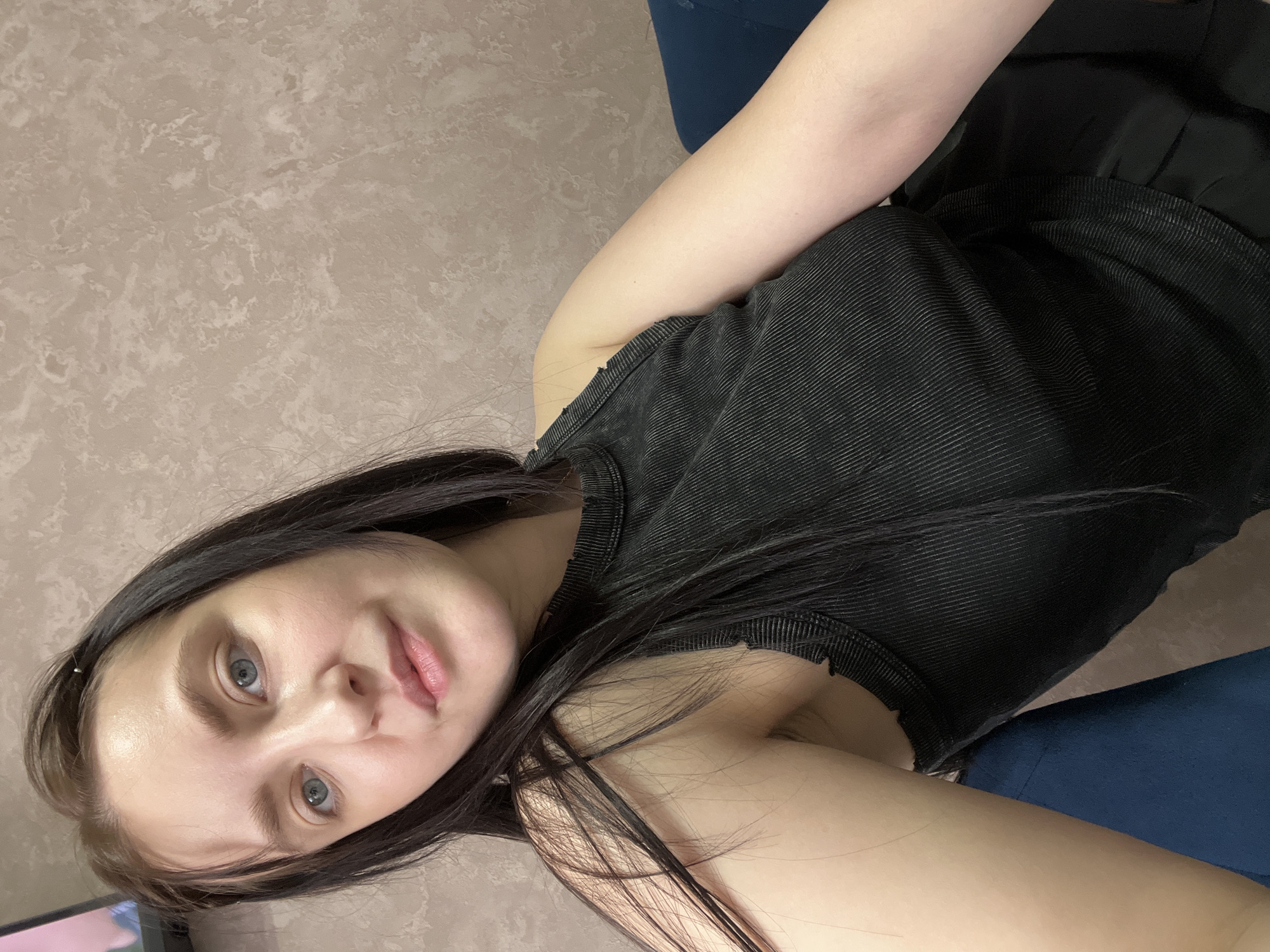 A young woman is photographed indoors against a backdrop of a textured beige wall. She is wearing a black tank top. Her dark hair falls across her shoulder and onto her chest. She has pale skin, light blue eyes, and pink lips. The woman is reclining on a dark blue surface. A partial view of a television screen is visible in the lower left corner. There is no indication of the location being Марковский, Russia.