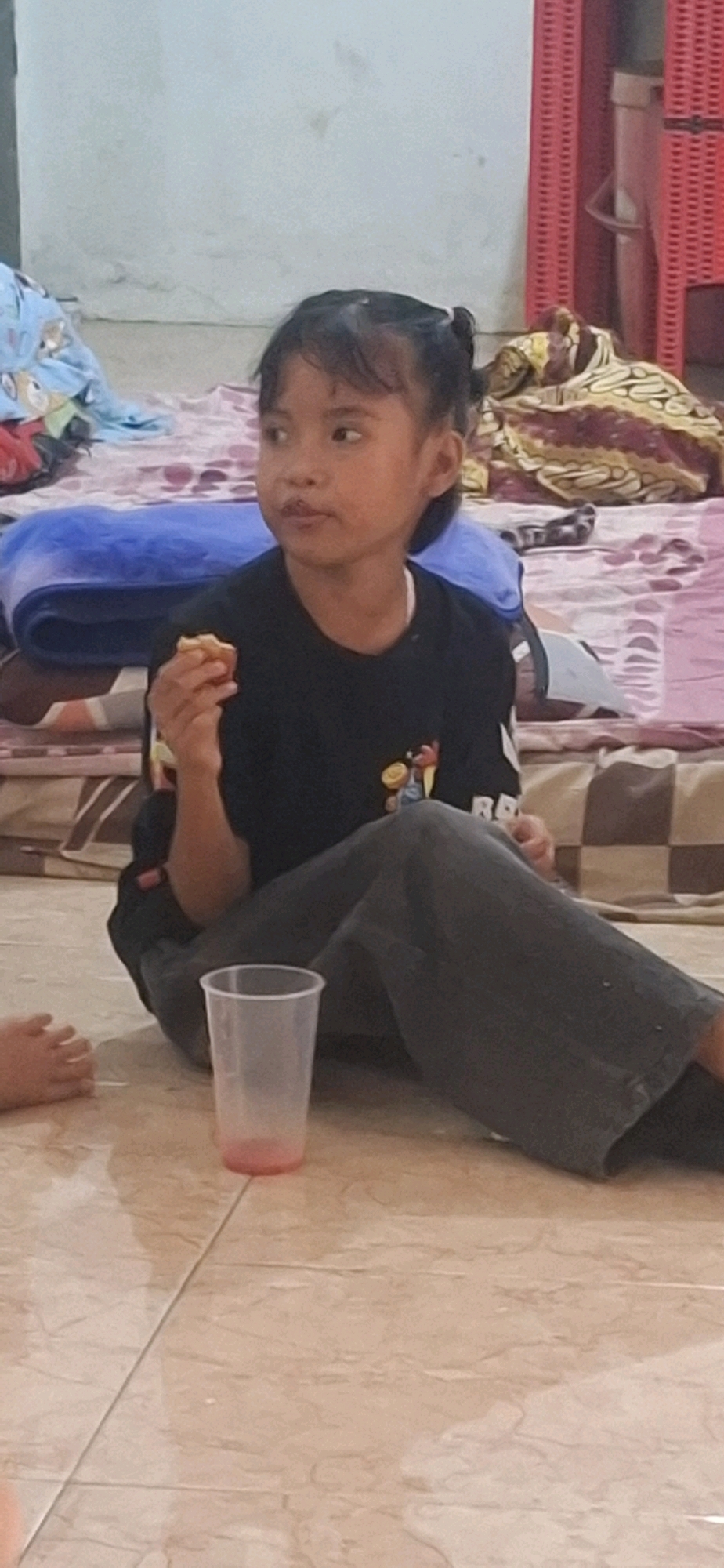 A young girl with dark hair tied in pigtails sits on the floor of a room, looking to her left. She is wearing a black t-shirt with a graphic print and dark grey pants. The girl is holding a small, light-colored object, possibly food, in her right hand near her mouth. In front of her, on the tiled floor, is a clear plastic cup with a pink liquid at the bottom. The background shows a cluttered area with folded blankets and what appears to be a red plastic chair or storage unit. The scene is set indoors, possibly in a residential area of Cibinong Hilir, Indonesia.