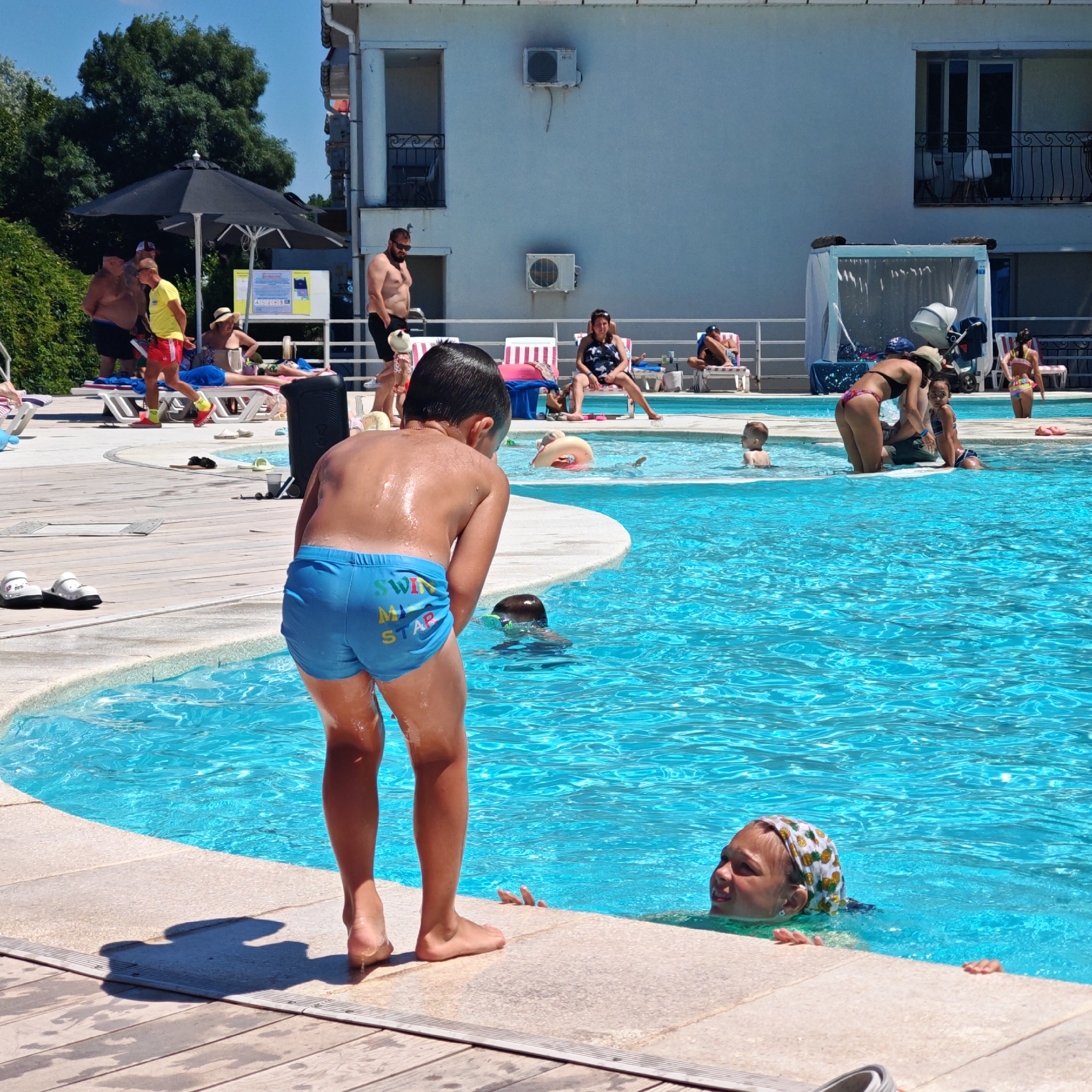 The image depicts a sunny day at an outdoor swimming pool, likely at a resort or hotel. In the foreground, a young boy wearing blue swim trunks with "SWIM MY STAR" printed on them stands on the pool deck, facing away from the camera and towards the water. His back is wet and glistening in the sunlight. Partially submerged in the turquoise water, to the right of the boy, is another child wearing a patterned headscarf. Their face is visible, looking upwards.
In the midground, several people are scattered around the pool area. A man in a black shirt and shorts stands on the left side of the pool, near a group of lounge chairs. Another man, wearing a yellow shirt and red shorts, is also on the deck, appearing to be an attendant or lifeguard. There are several lounge chairs with towels and a black speaker system on the pool deck. Further back, a woman in a black and white patterned swimsuit is seated on a lounge chair. In the background, a white building with balconies and air conditioning units is visible. The overall environment is bright and appears to be a recreational area.