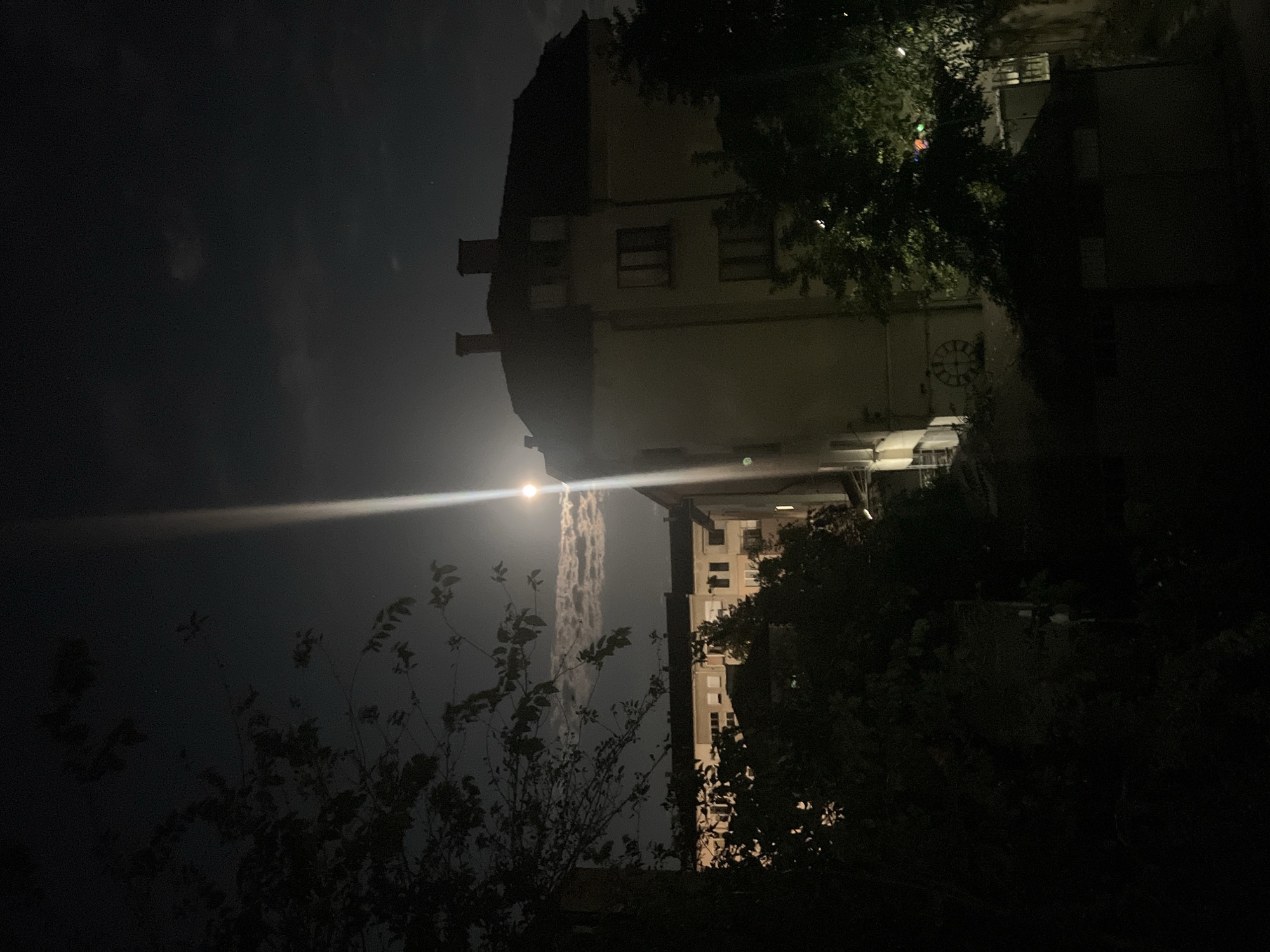The image depicts a nighttime scene in Blagoevgrad, Bulgaria, featuring buildings illuminated by moonlight. In the foreground, trees with dark foliage partially obscure the view. Behind the trees, a building with multiple windows and dormer windows on its roof is visible, bathed in a strong shaft of moonlight that also creates a lens flare effect. Another, taller building with a lighter facade and numerous windows is visible to the left of the main building, also illuminated by the moon. The sky is dark, with some patchy clouds and a bright moon visible. Part of a building with a circular clock face is visible on the right side of the image, partially obscured by trees. The overall impression is one of a quiet, nocturnal urban environment.