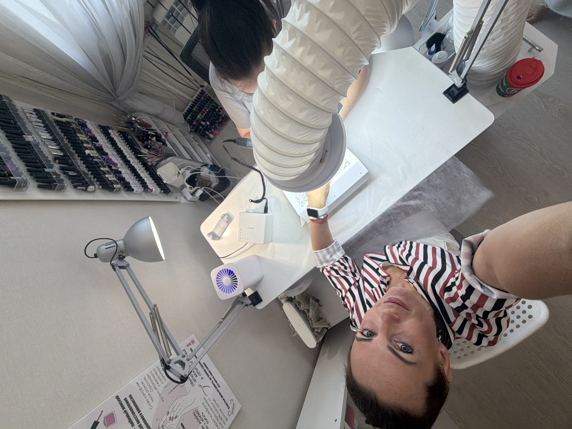 The image depicts a top-down view of a nail salon located in Birobidzhan, Russia. A woman with dark hair, wearing a striped shirt, is seated and looking at the camera. Her right arm is extended towards a white table where another person, whose face is not visible, is working on nail services. The setup includes a nail lamp, a dust collector, various bottles of nail polish displayed on wall-mounted shelves, and other nail art supplies. The lighting appears to be artificial, originating from the overhead lamp. The environment suggests a professional workspace dedicated to nail care.