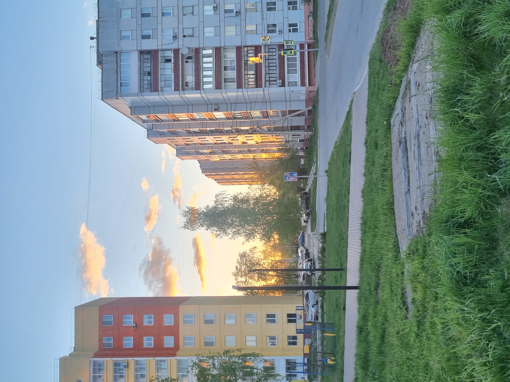 The image depicts a street scene in Chaikovsky, Russia at sunset. Two multi-story apartment buildings stand opposite each other, with a street and sidewalk between them. The building on the left has a yellow and red facade, while the one on the right is gray. The setting sun casts a golden glow on the buildings and the sky, visible through the trees between them. Several cars are parked along the street, and a playground with colorful equipment is visible near the building on the left. The sidewalk is paved with bricks, and tall green grass grows along the edge of the sidewalk. A street light is visible near the buildings.