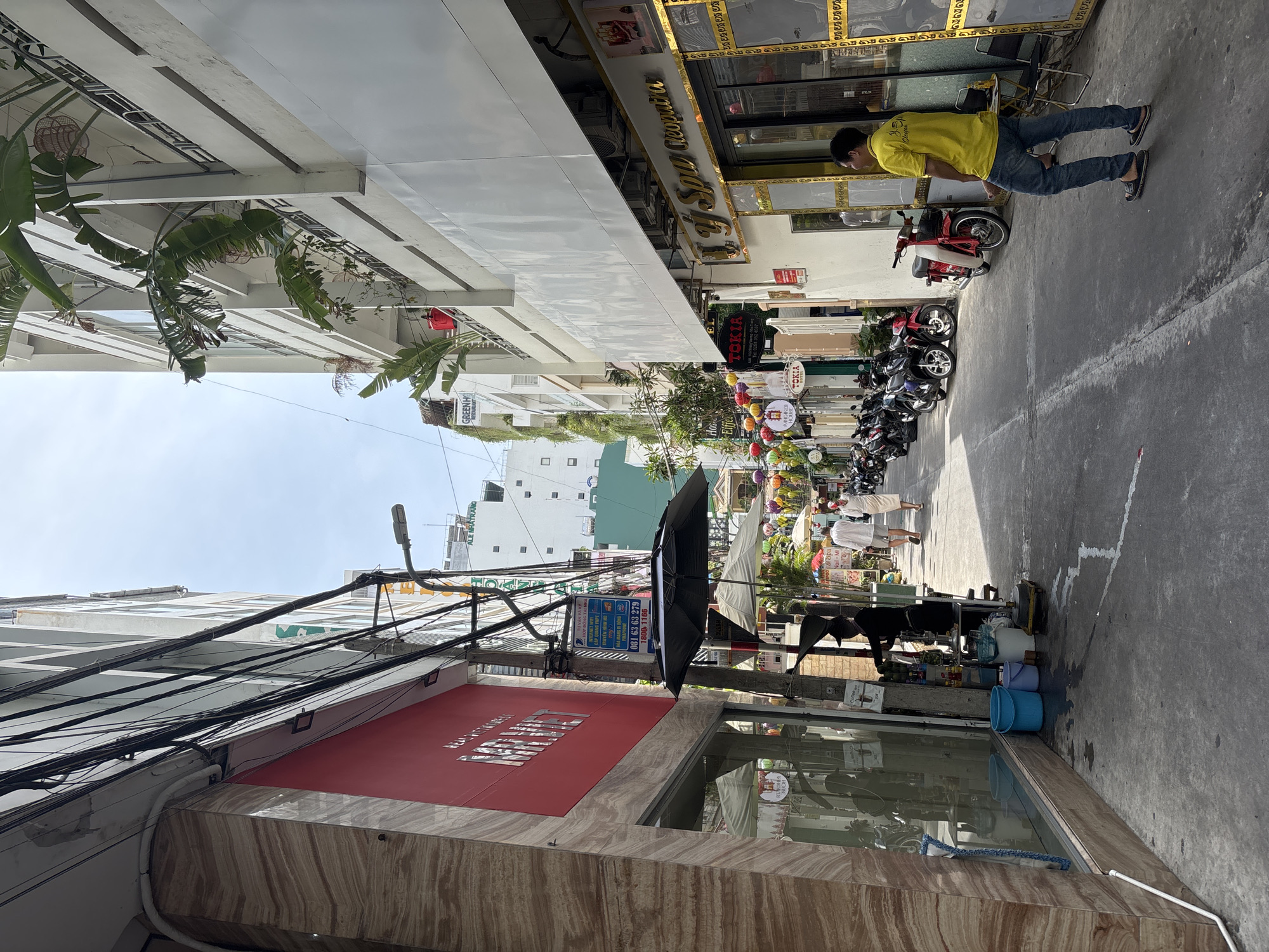 The image depicts a narrow street scene in Nha Trang, Vietnam. On the left, a building features a red facade with textured marble accents. A glass storefront displays reflections of the street and people inside. A man in a dark outfit stands at a cart near the storefront, seemingly preparing drinks. Several buckets are visible on the sidewalk.
The street is lined with parked motorcycles, primarily scooters. On the right side of the street, a building is adorned with a sign for "U LY SPA Cleopatra" in gold lettering. A man in a yellow t-shirt and blue jeans stands on the sidewalk facing the spa entrance.
Further down the street, colorful lanterns hang overhead, adding a festive atmosphere. People are walking along the street, including two individuals in white garments. Buildings on both sides of the street are multi-story structures with balconies, some featuring plants. Overhead, a dense network of electrical wires is visible against the sky. The overall scene suggests a bustling urban environment with commercial activity.
