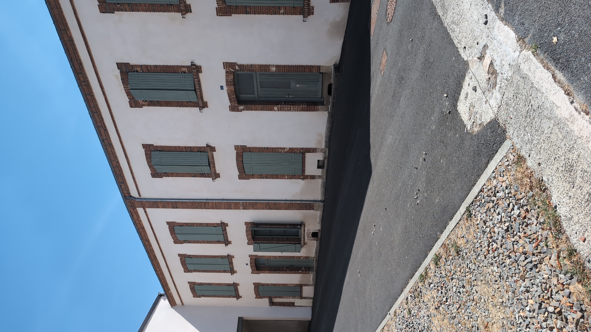 The image shows a close-up view of a building's facade and the adjacent street in the city of Cholet, France. The building has a white plaster exterior with red brick accents around the windows and at the roofline. Several windows with green shutters are visible, as well as a grey double door. The street in front of the building is paved with asphalt and bordered by a gravel area. The sky is a clear, bright blue. The scene appears to be a sunny day.