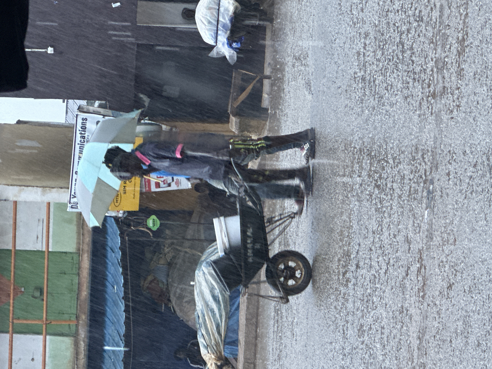 In the city of Bukuru, Nigeria, a heavy downpour is evident as rain streaks down, creating a wet and reflective surface on the street. Two individuals are visible, walking side-by-side. The person in the foreground, on the right, is pushing a wheelbarrow. This wheelbarrow is dark-colored and contains a white bucket and other items covered with a tarp. The person pushing the wheelbarrow is wearing a dark shirt with pink trim on the collar and sleeves, and dark pants with bright green and yellow striped accents. They are also wearing dark shoes. To their left, another person is walking and holding a light blue and grey umbrella over both of them. This person is wearing a blue shirt and dark pants. In the background, a building with a blue awning is visible, along with signs advertising "MTN SIM R" and "Communications". The overall scene depicts people navigating the adverse weather conditions in a city setting.