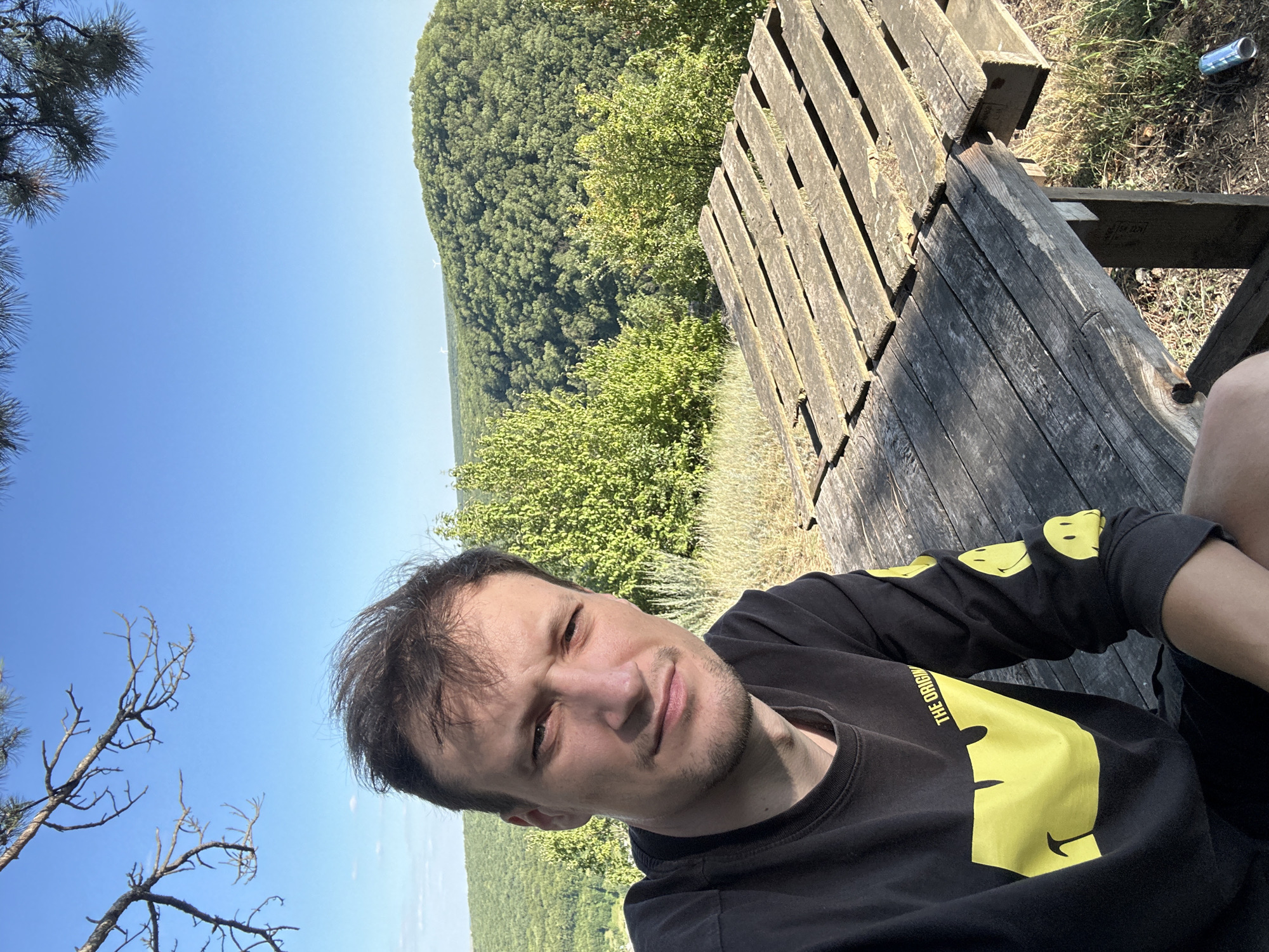 This is a selfie taken by a man sitting on a wooden bench outdoors. The man is wearing a black long-sleeved shirt with yellow smiley face graphics on the sleeves and chest, and text that appears to say "THE ORIGINAL". He has short, dark hair and is looking towards the camera with a slight smile. The background shows a lush, green forest stretching out to the horizon under a clear blue sky. The bench is made of weathered wooden planks and appears to be part of a picnic table or seating area. In the upper right corner of the image, a metallic can is visible on the ground next to the bench. The scene suggests a bright, sunny day, likely in the late morning or early afternoon, given the strong sunlight and clear sky. The overall setting is a natural, scenic location, possibly a park or a viewpoint overlooking a forest.