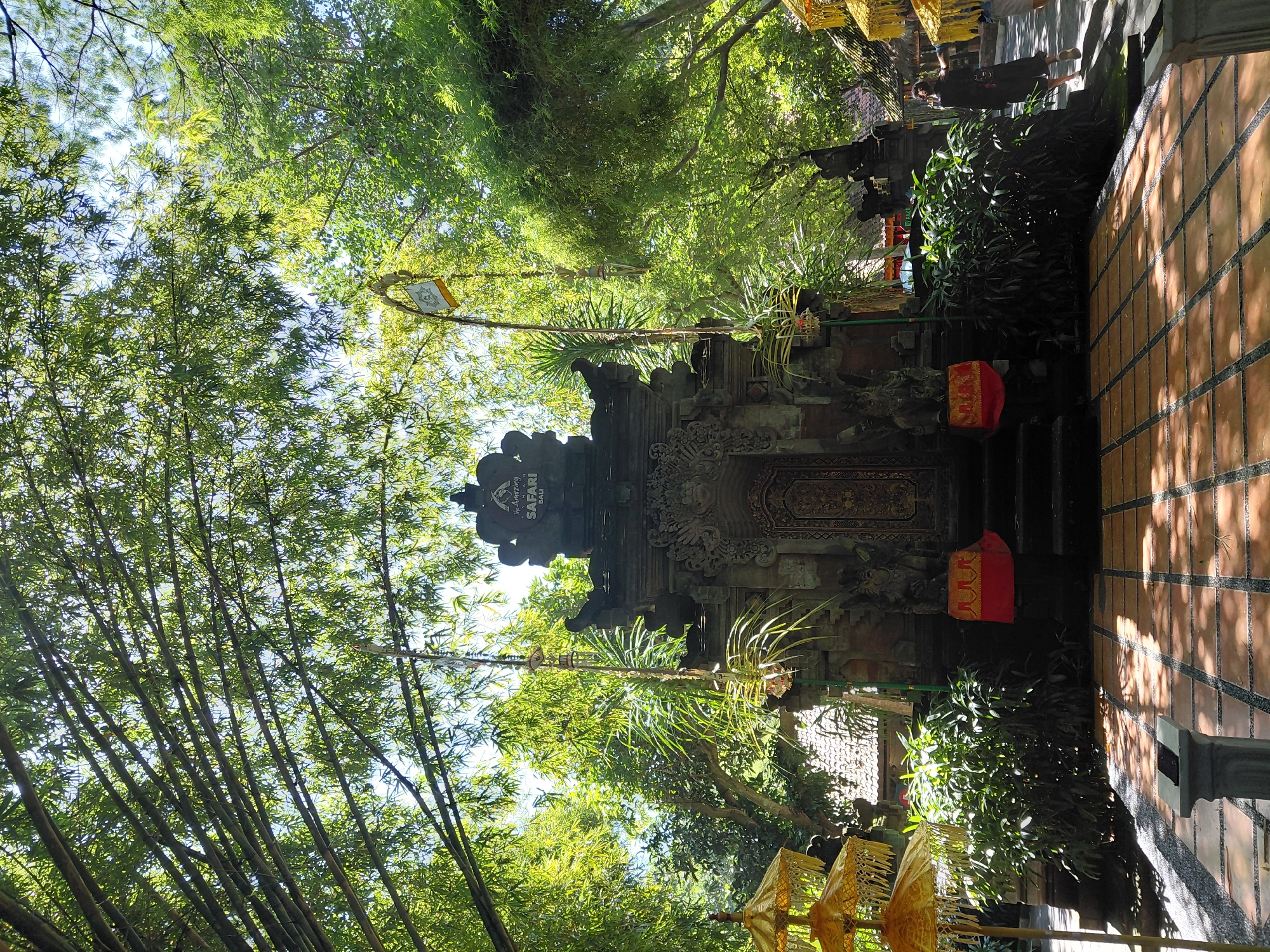 The image depicts a Balinese temple structure at The Amazing Safari Bali, located in Blahbatuh, Indonesia. The temple is ornately carved from stone and features a traditional tiered roof. A detailed, golden doorway is the central focus of the temple facade. Flanking the entrance are stone statues of guardians. Red cloths are draped over the lower steps. Lush green foliage, including bamboo trees, surrounds the temple, with sunlight filtering through the leaves. Yellow ceremonial umbrellas are visible on the left and right sides of the scene, adding to the festive atmosphere. A pathway paved with terracotta tiles leads to the temple. In the background, a person wearing a dark outfit walks away from the camera, and another person in lighter clothing is partially visible. The environment is outdoors, under a bright sky.