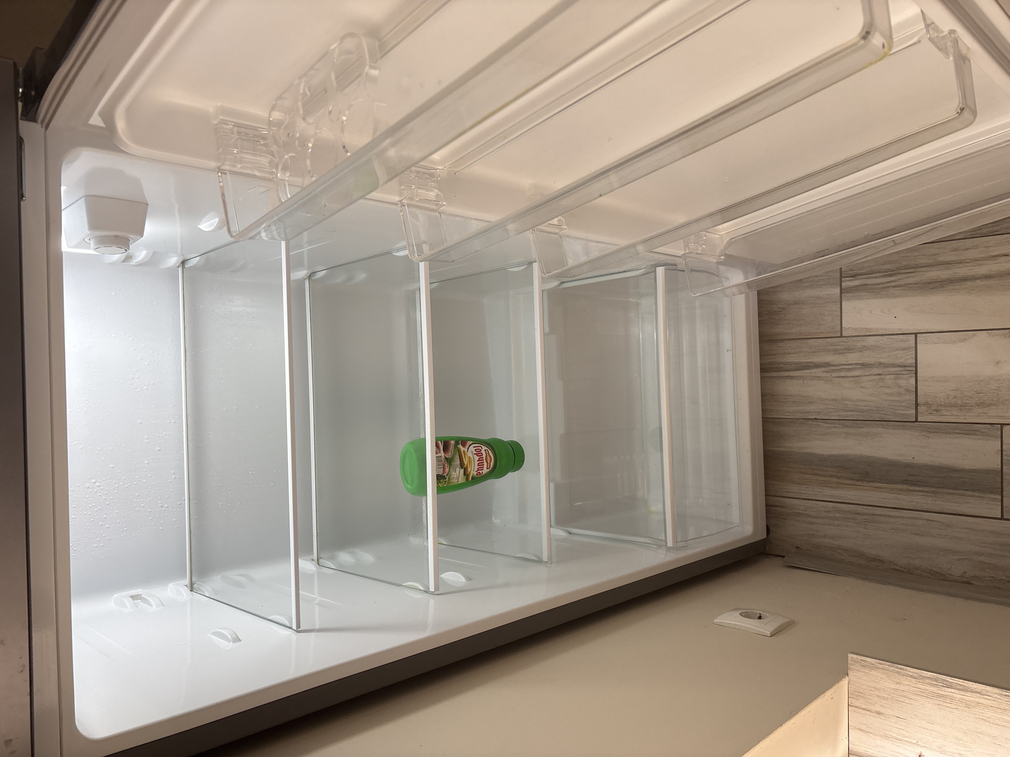 The interior of an open refrigerator is shown. The refrigerator is largely empty, save for a single green bottle of "Горчица" (Mustard) lying horizontally in one of the compartments. The refrigerator is white and silver, with clear plastic dividers creating several compartments. The back wall has condensation droplets on it. Outside the refrigerator, the countertop and a section of tiled wall can be seen. The location is likely indoors, potentially in a residential or commercial kitchen, based on the countertop and tiled wall. There is no explicit indication to suggest the scene is in Sofia, Bulgaria.