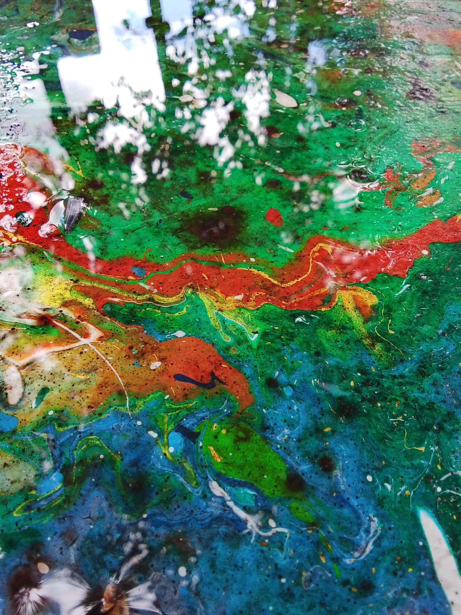 The image displays a close-up, abstract view of paint poured on a surface, likely a table or canvas, with a reflective, watery element. The dominant colors are vibrant greens, blues, reds, and yellows, swirling and blending into each other with organic, fluid patterns. There are also flecks of white and darker specks scattered throughout. The surface appears wet, with reflections of what could be a bright sky and foliage, suggesting an outdoor or well-lit environment. There are some small bubbles visible in the liquid. The lower portion of the image shows a blurred, possibly organic object submerged in the liquid, with white, feathery protrusions. There are no discernible people or specific city landmarks of 1-Go Maya, Russia.