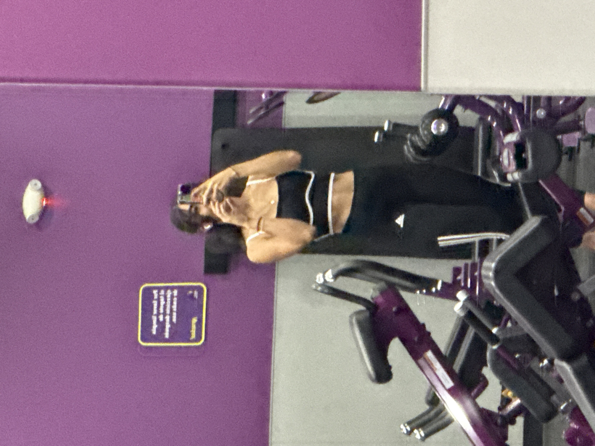 The image shows a mirrored reflection of a woman in a gym, possibly at a Planet Fitness in Puebla, Mexico, given the gym's typical color scheme. The woman is holding a phone up, likely taking a picture of herself in the mirror. She is wearing a black sports bra with white trim and black leggings with an Adidas logo. Several pieces of purple and black gym equipment (possibly exercise bikes) are visible. The gym wall is a bright shade of purple with a small informational sign and a white emergency light attached to it.